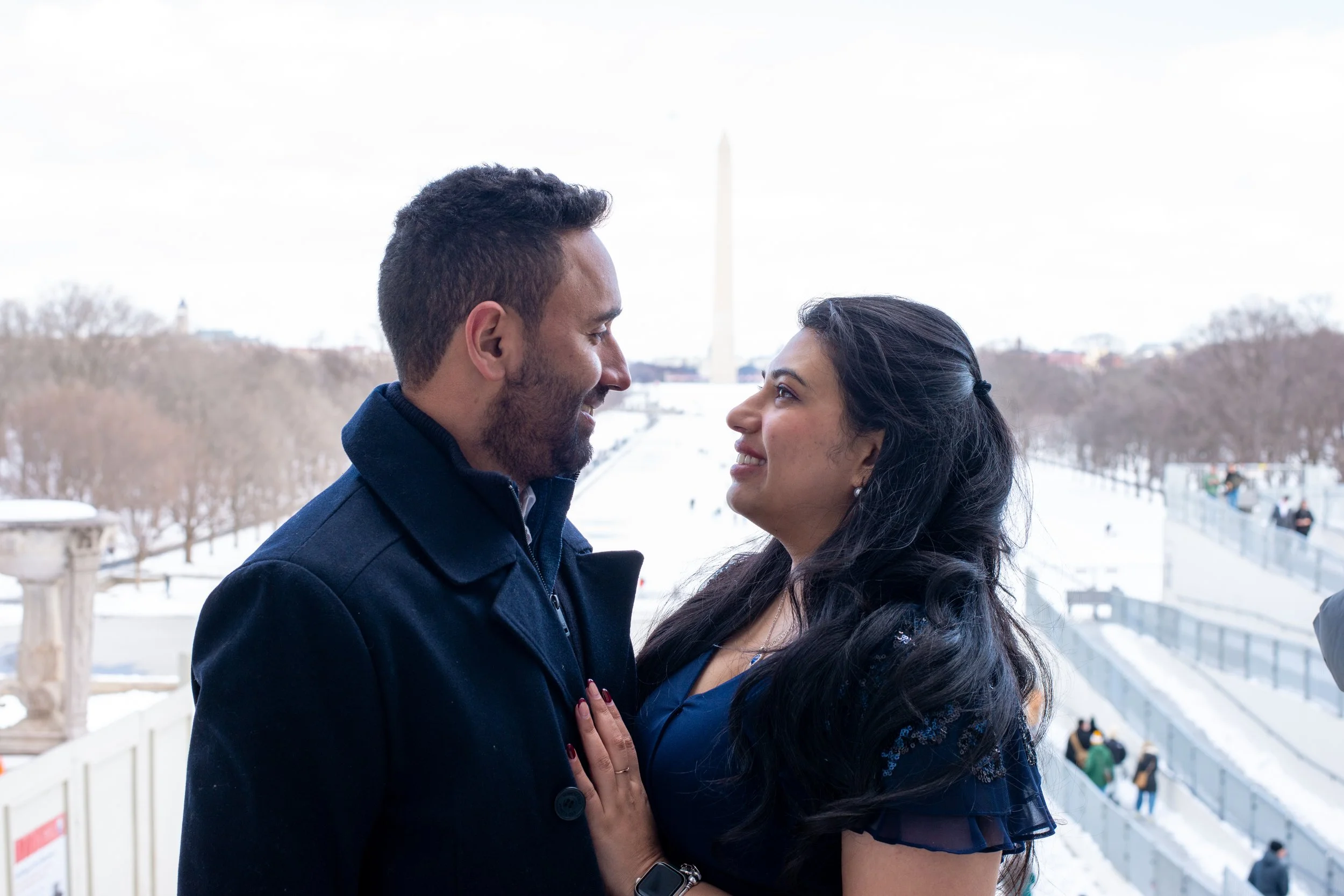 A man and woman face to face outdoors in winter, smiling and holding hands, with the Washington Monument in the background.
