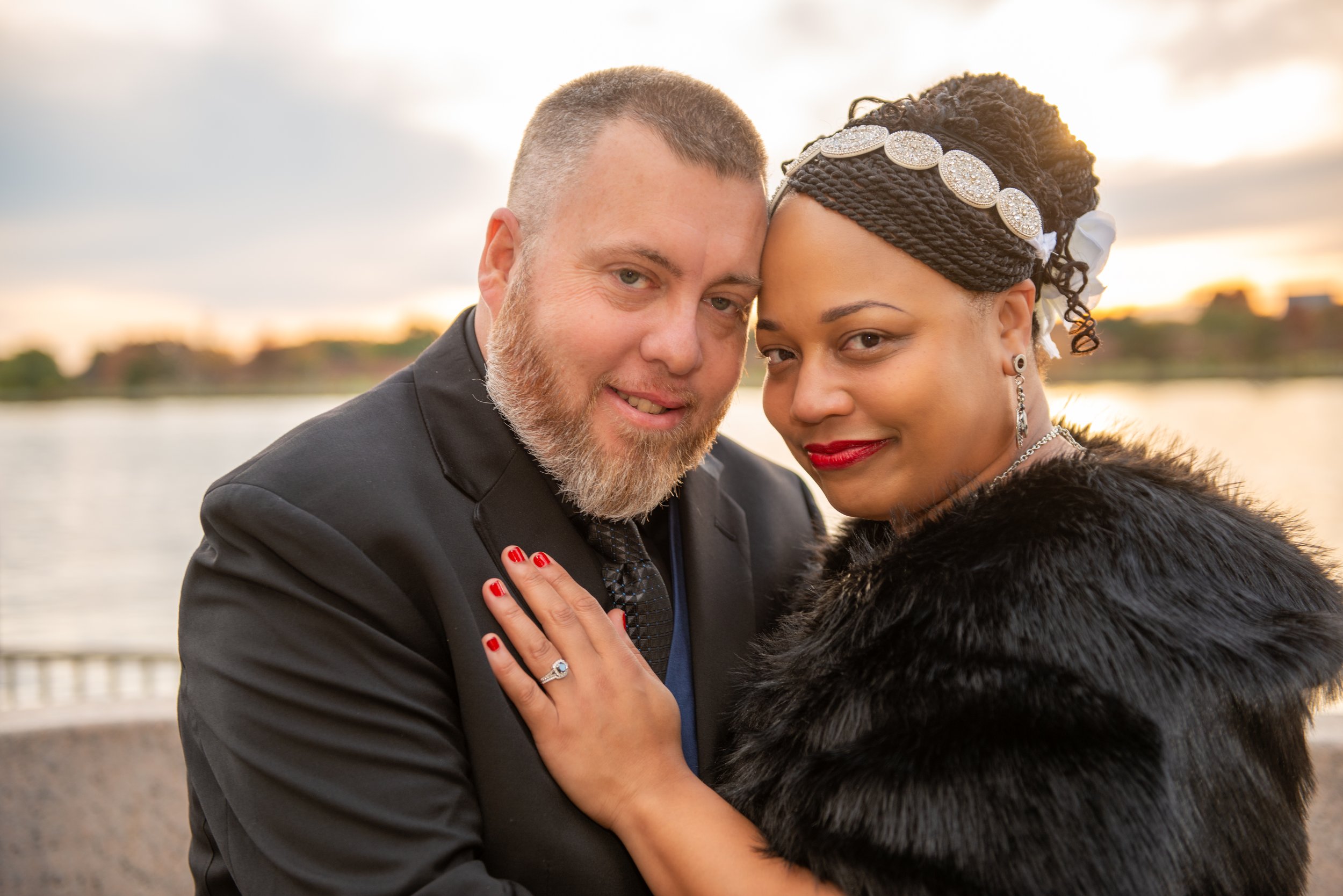 A couple dressed elegantly, embracing near a body of water at sunset, with the man wearing a black suit and the woman wearing a black fur coat and decorative headwrap.