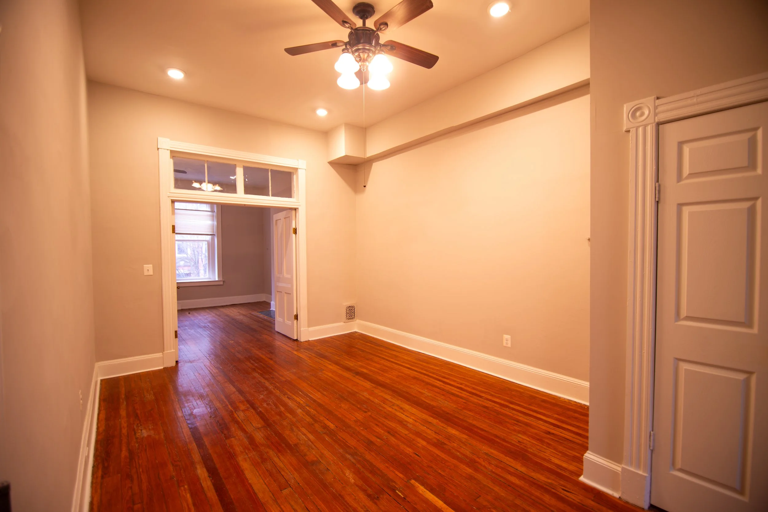Empty room with hardwood floors, cream-colored walls, a ceiling fan with lights, recessed ceiling lights, a doorway leading to another room, and a window with a shade.
