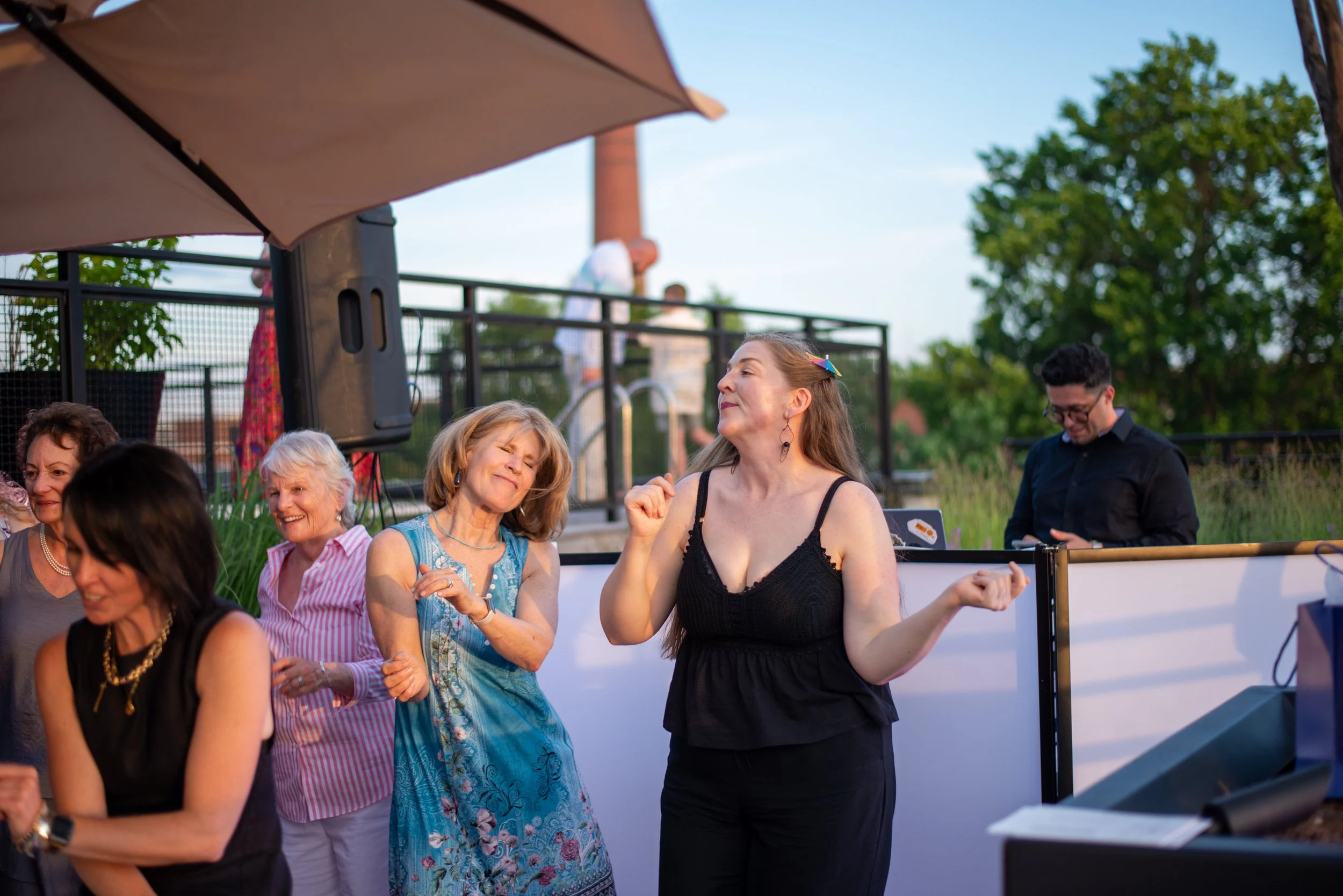 Group of people at an outdoor event with some dancing or celebrating, people smiling and enjoying the moment, trees and a building in the background.