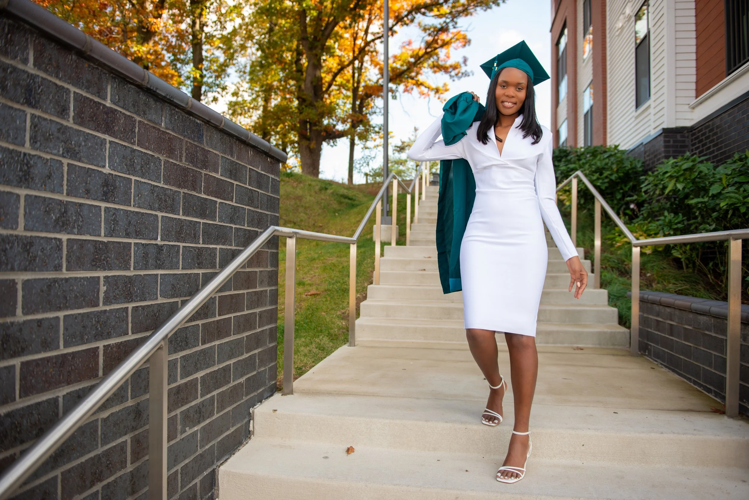 A woman in a white dress and high heels walking down outdoor steps, carrying a graduation gown over her shoulder, with autumn trees and residential buildings in the background.