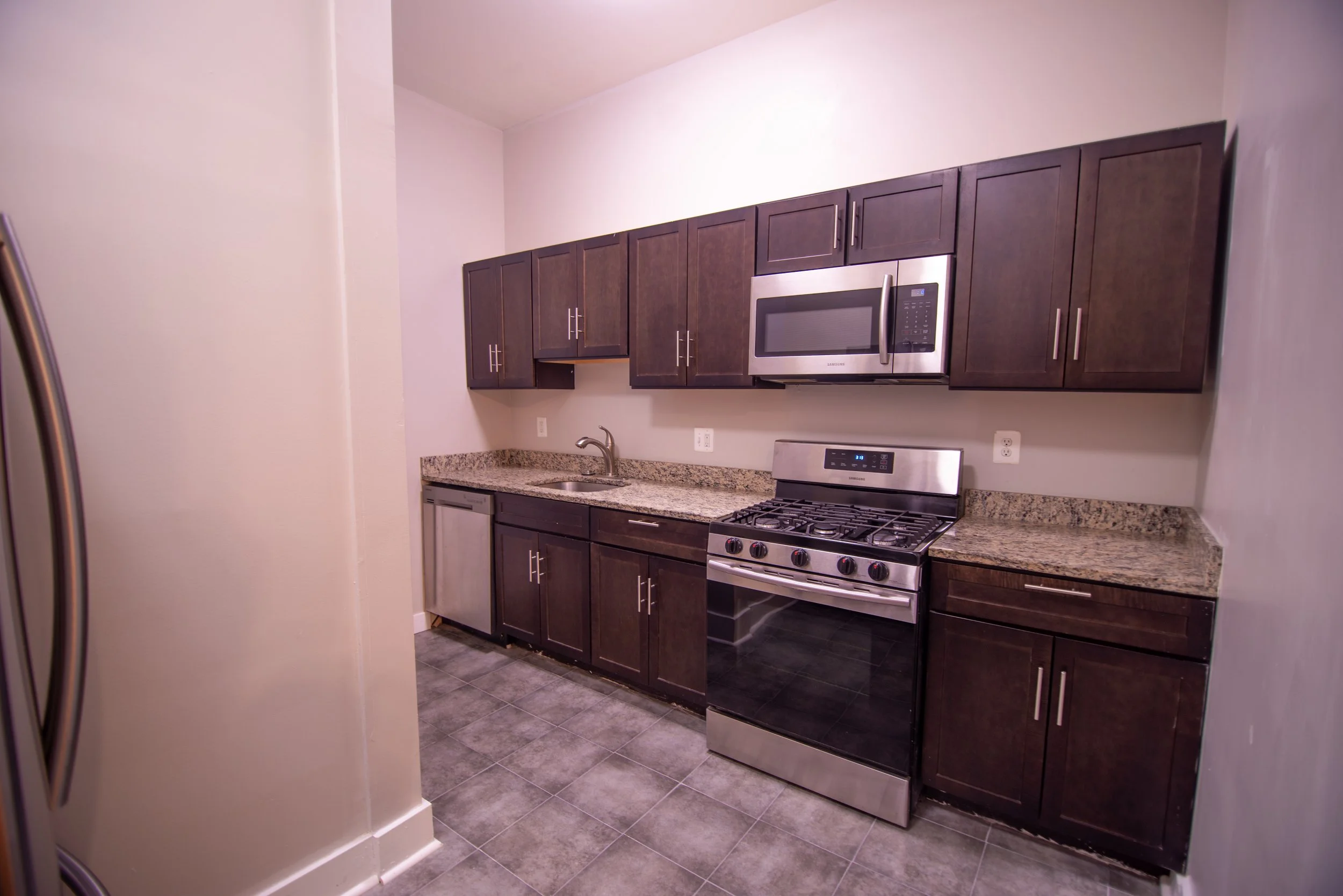 A kitchen with dark wooden cabinets, a stainless steel microwave, a stove, a granite countertop, and a small refrigerator, with gray tiled flooring and white walls.