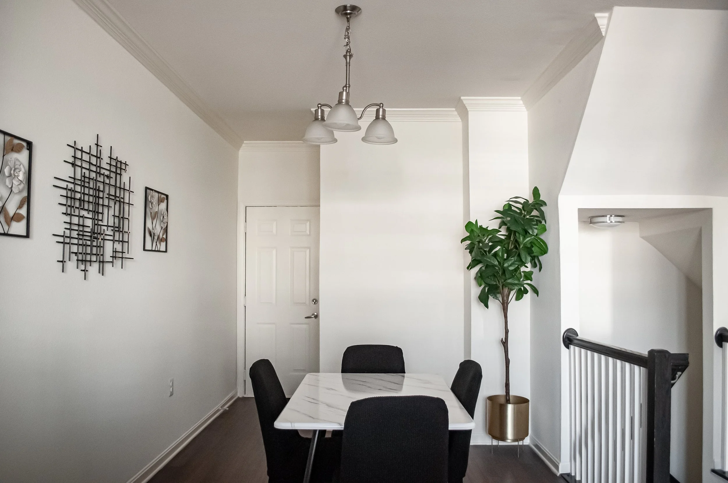 A dining room with a white marble table surrounded by five black chairs, a green potted plant in a gold pot, artwork on the wall, and a white door in the background. A modern light fixture hangs from the ceiling.