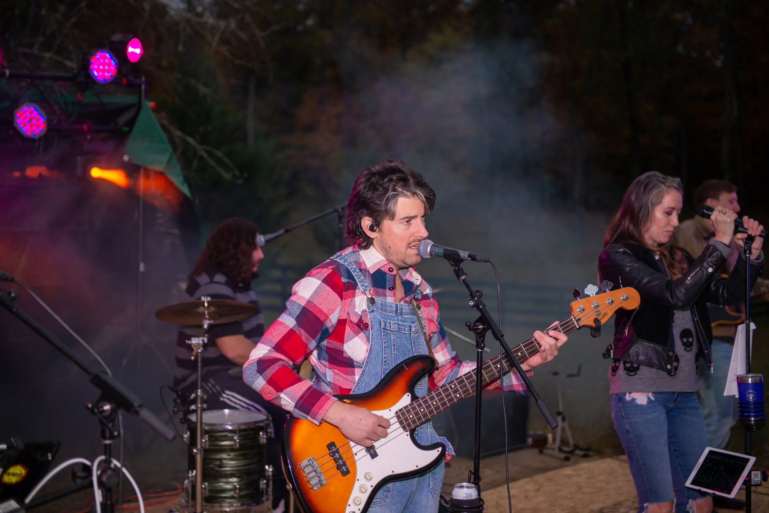 A group of musicians performing outdoors at night, including a man playing an electric bass guitar and singing into a microphone, with other band members and musical equipment visible in the background.