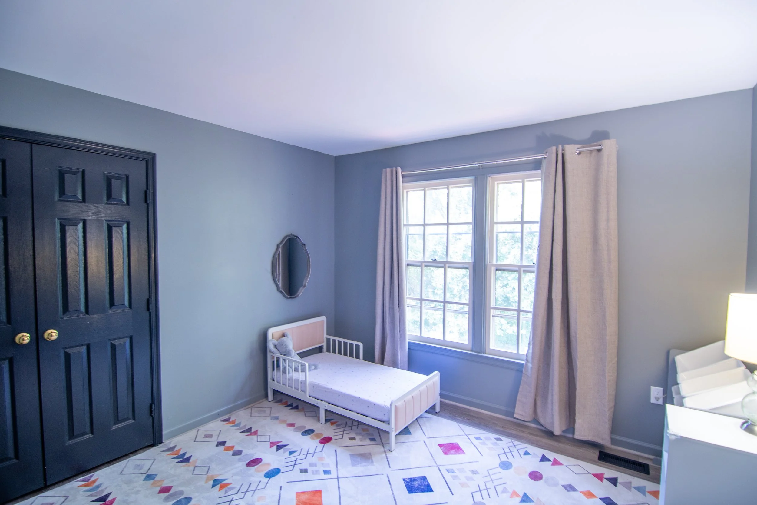 A corner of a room with a white crib, a large window with beige curtains, a blue wall, a black closet door, and a colorful geometric patterned rug.
