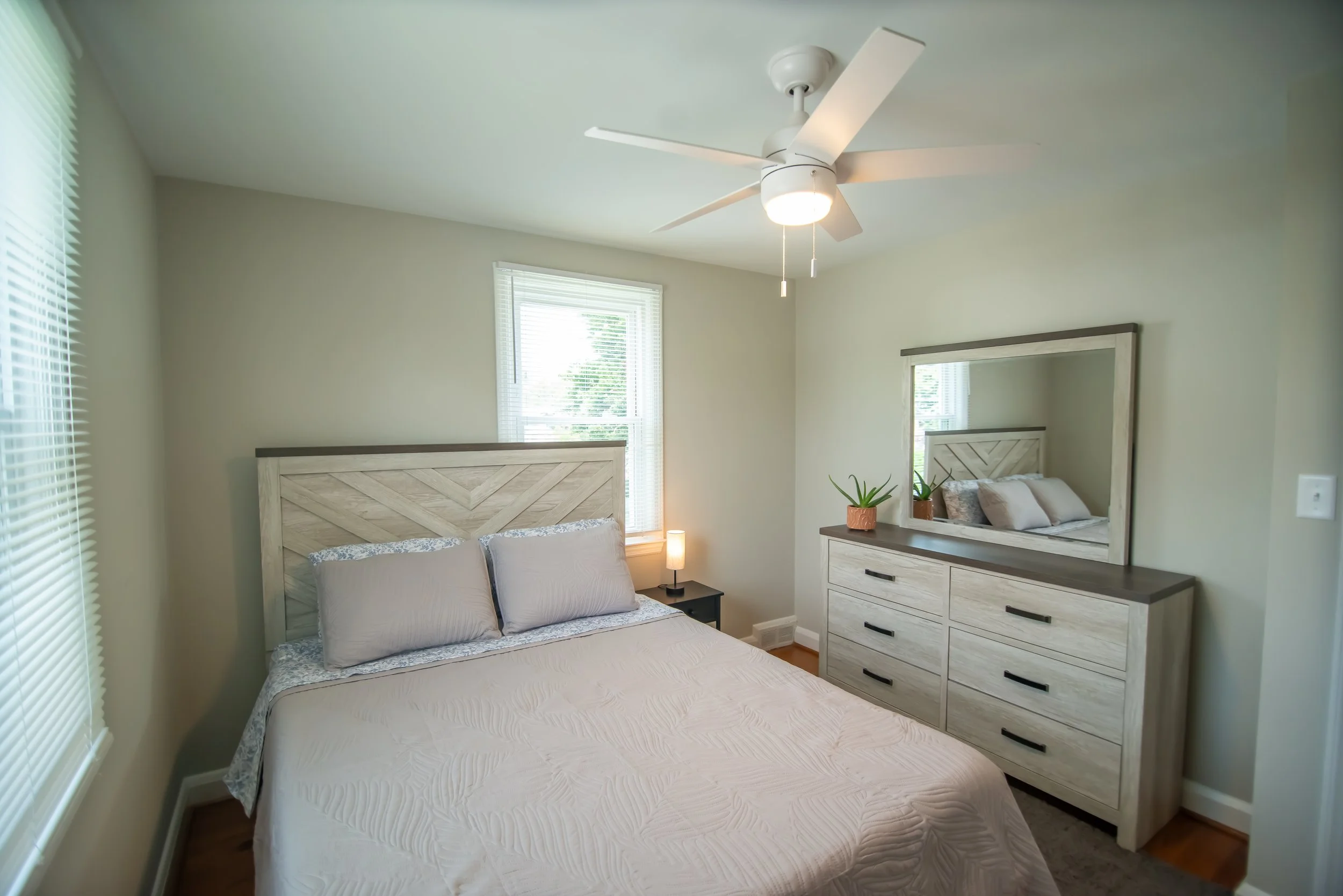 A bedroom with a white bed, nightstand, and dresser, with a large mirror and a ceiling fan, illuminated by sunlight through windows with blinds.