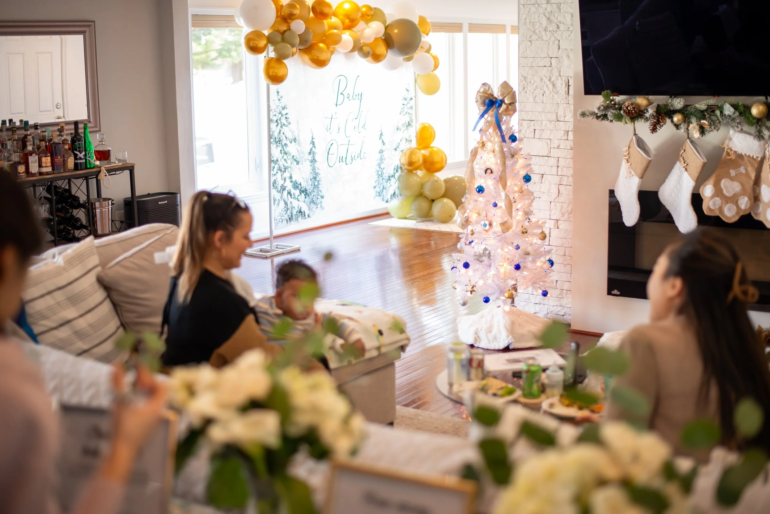 Indoor Christmas gathering with holiday decorations, Christmas stockings hanging on the fireplace, decorated Christmas tree with blue and gold ornaments, large gold and white balloons, and a snowy outdoor scene visible through the window.