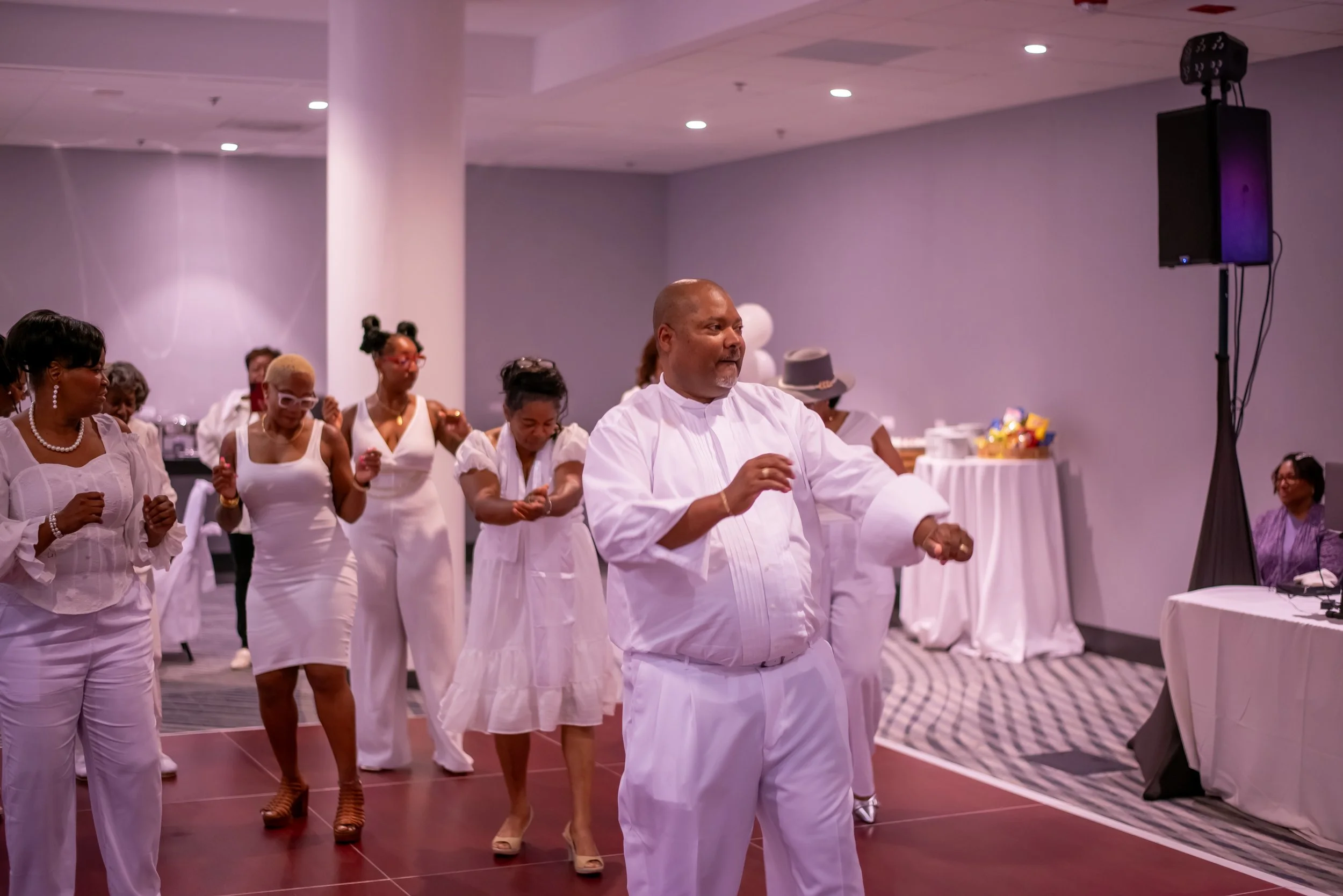 Group of people dancing in white clothing at an indoor event, with some wearing accessories like glasses and hats, and a large speaker to the right side of the image.