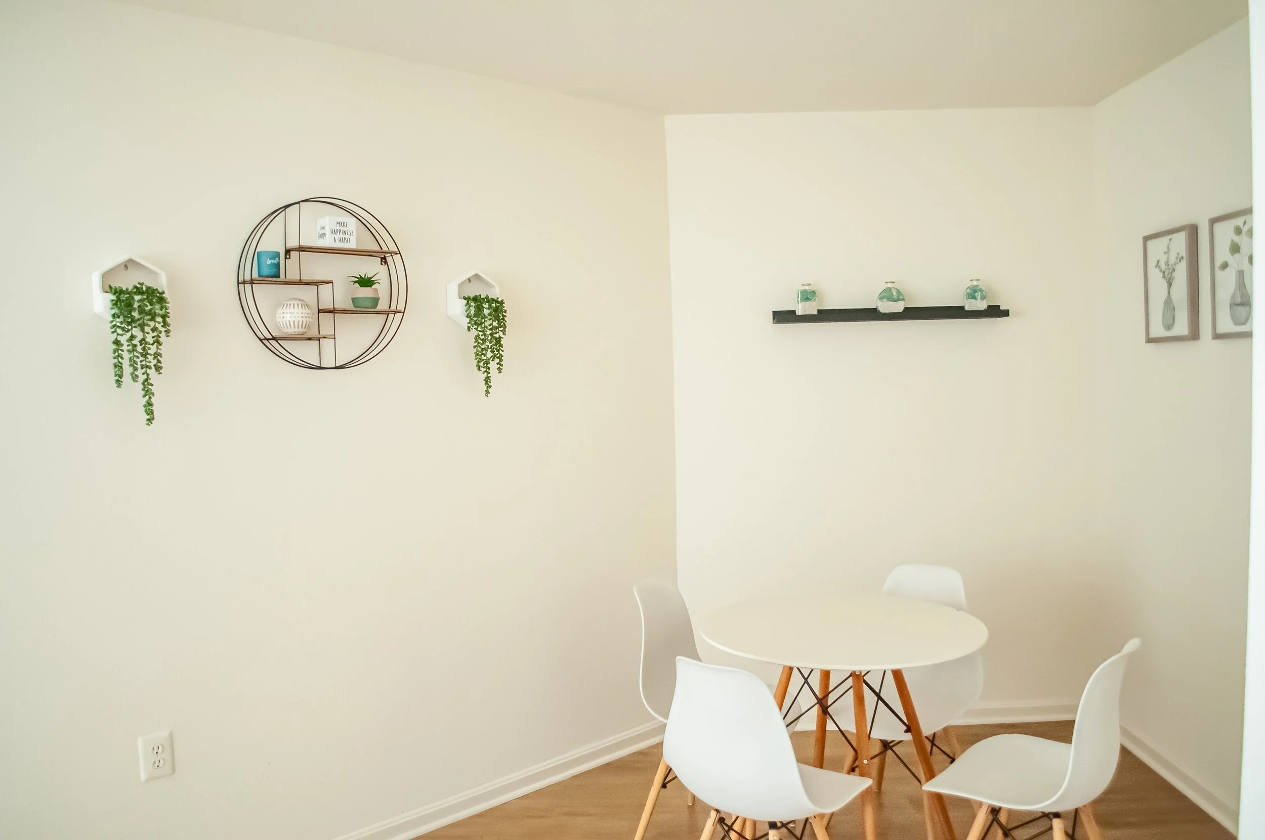 A minimalist dining area with a white round table, white chairs, a black shelf with three small decorative jars, and wall-mounted shelves with plants and decorations on a light-colored wall.
