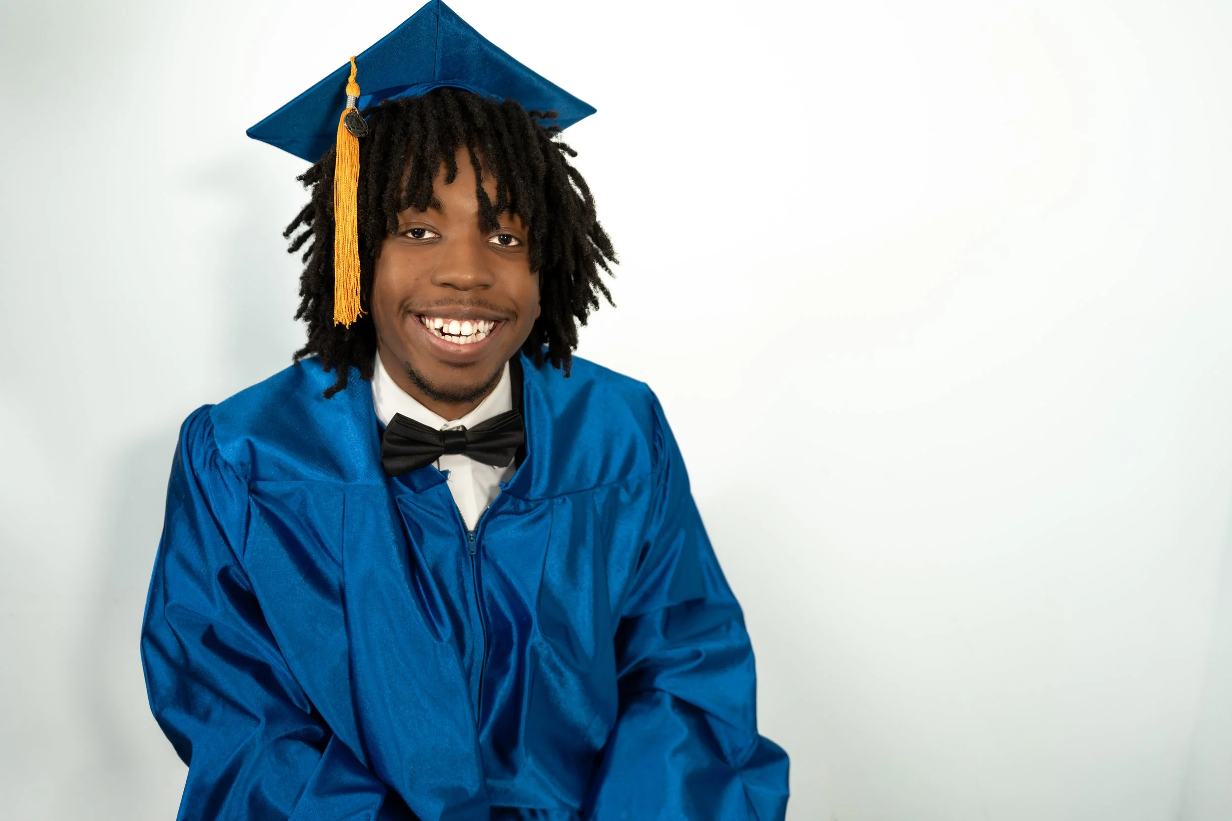 Young man wearing a blue graduation gown and cap with a yellow tassel, smiling, against a plain white background.