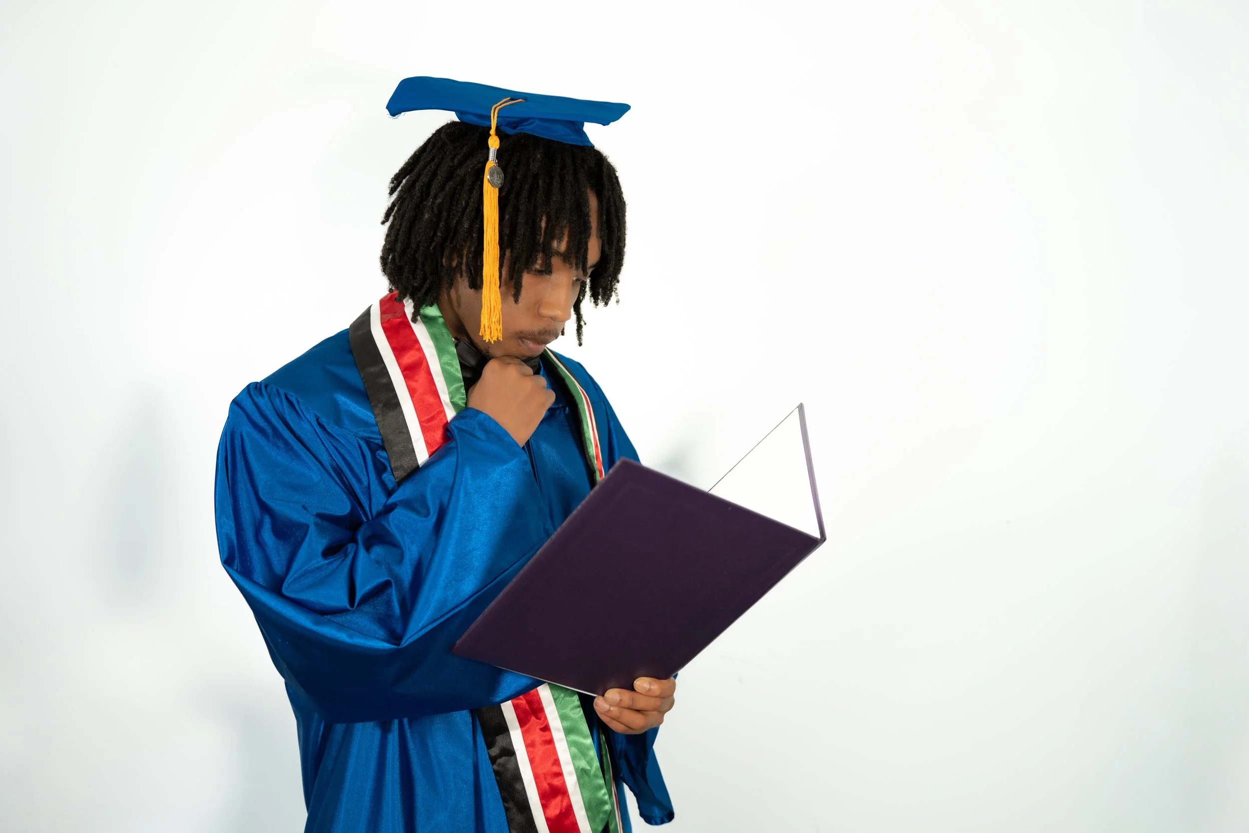 A young man in a blue graduation gown and cap holding a diploma while standing against a plain white background.