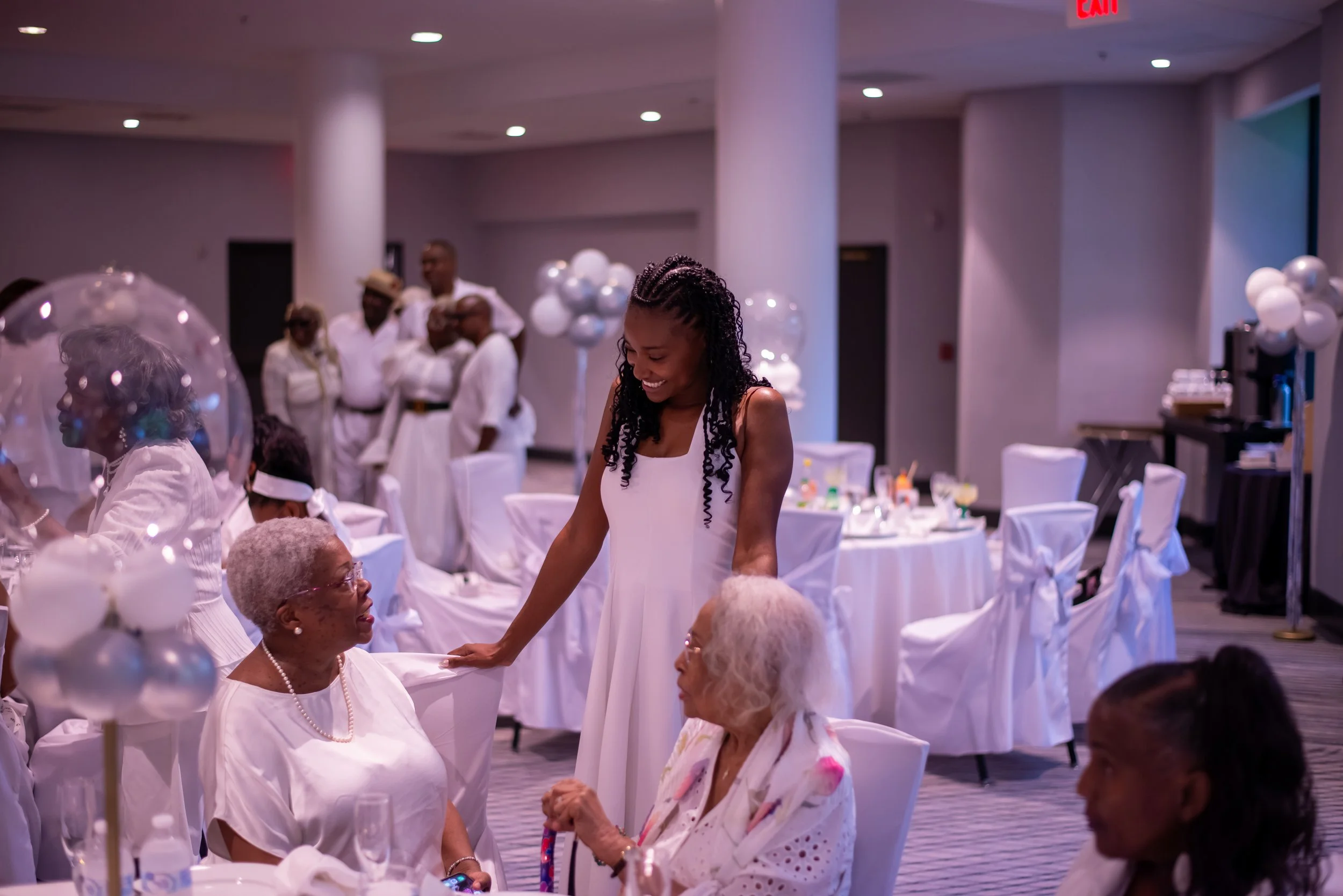 A young woman with braided hair smiling and talking to elderly women in a white dress at a celebration event with white decorations and balloons.