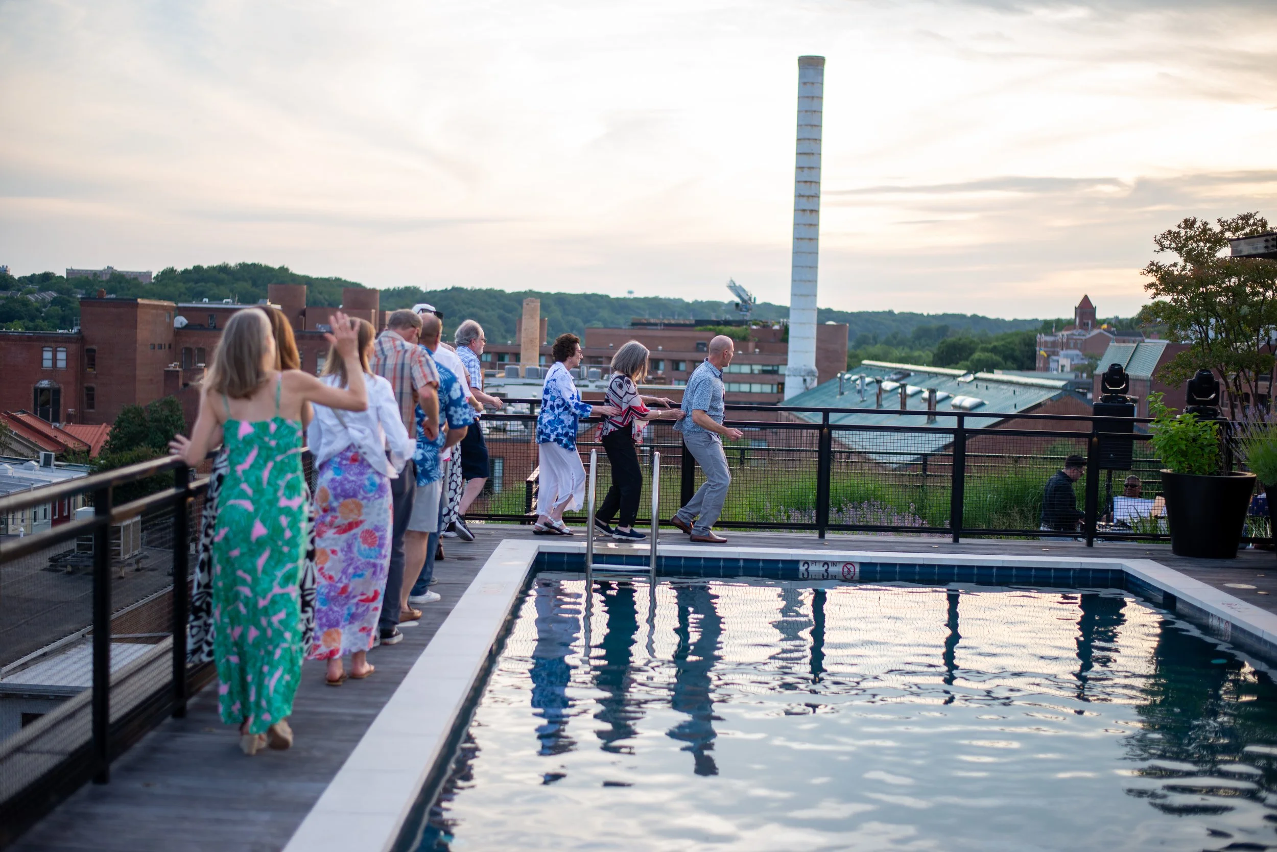 Group of people dressed in colorful clothing walking in a line beside a swimming pool on a rooftop at sunset. City buildings and greenery are visible in the background.