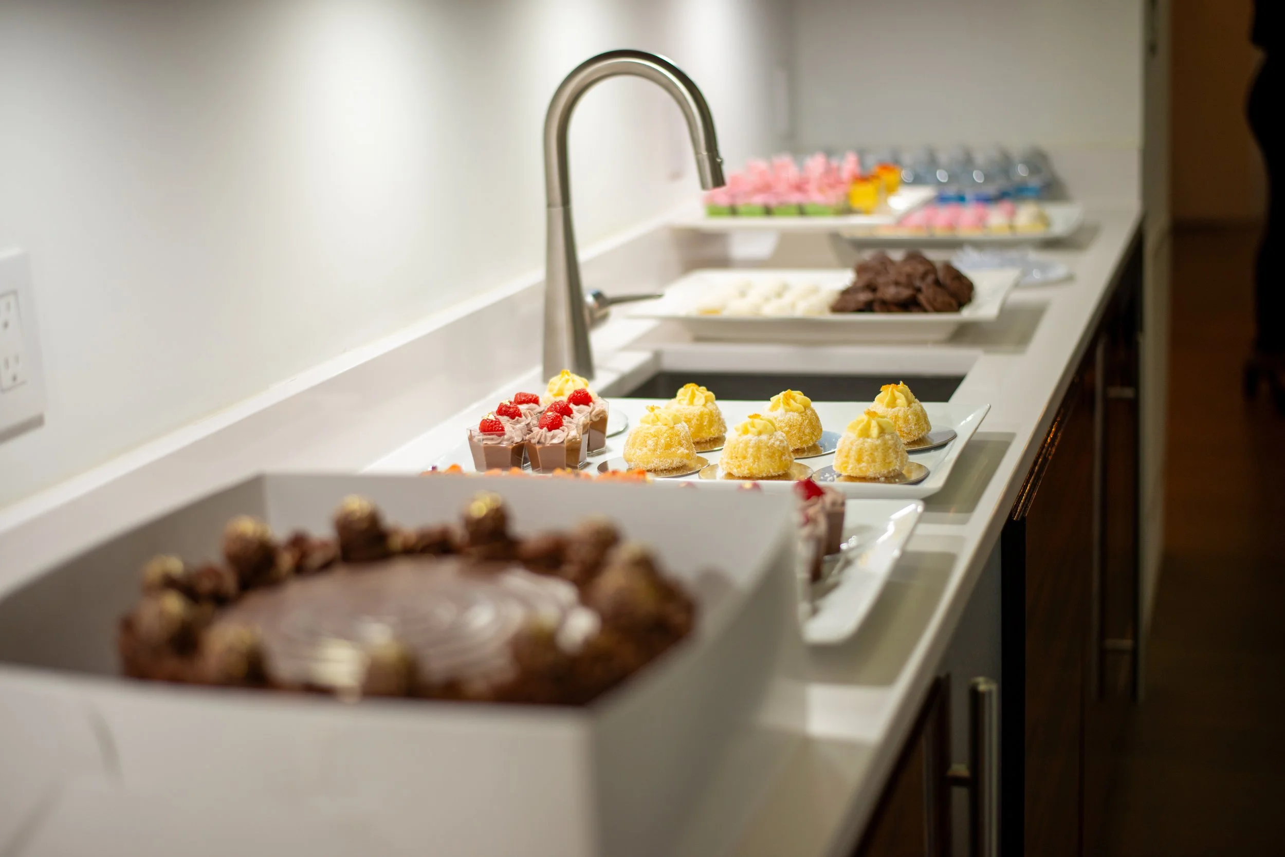 A row of assorted desserts on white trays, including cupcakes with pink flowers, yellow éclairs, and chocolate treats, on a kitchen counter with a sink and faucet, with bottles of water and pink decorations in the background.