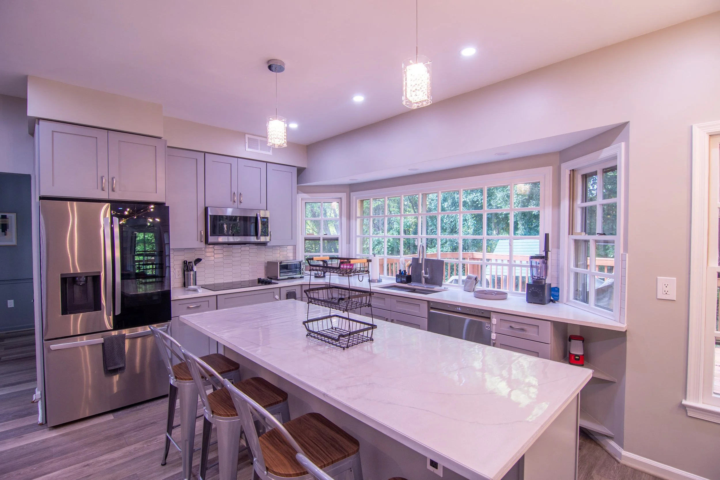 A modern kitchen with white cabinets and a large white island. Stainless steel refrigerator and microwave, large windows with a view of greenery, and pendant lights hanging from the ceiling.