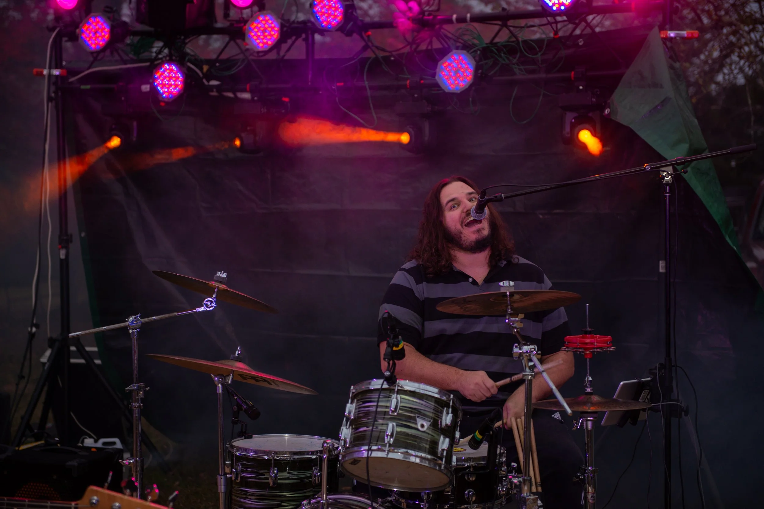 A musician with long curly hair and a beard playing drums on stage, singing into a microphone, with colorful stage lighting above him.
