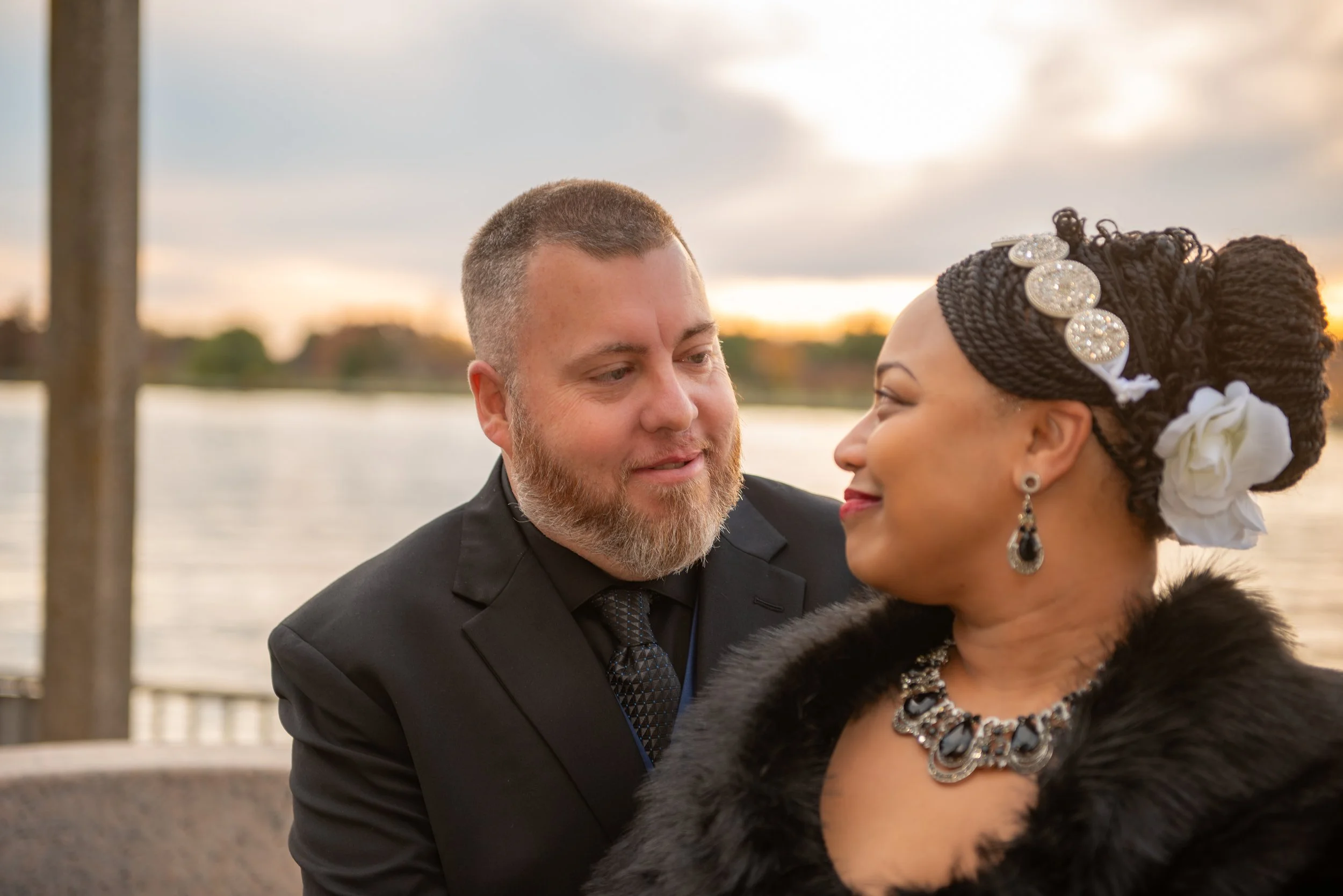 A couple dressed elegantly with the man in a suit and the woman wearing jewelry and a black head wrap, standing near a body of water during sunset.
