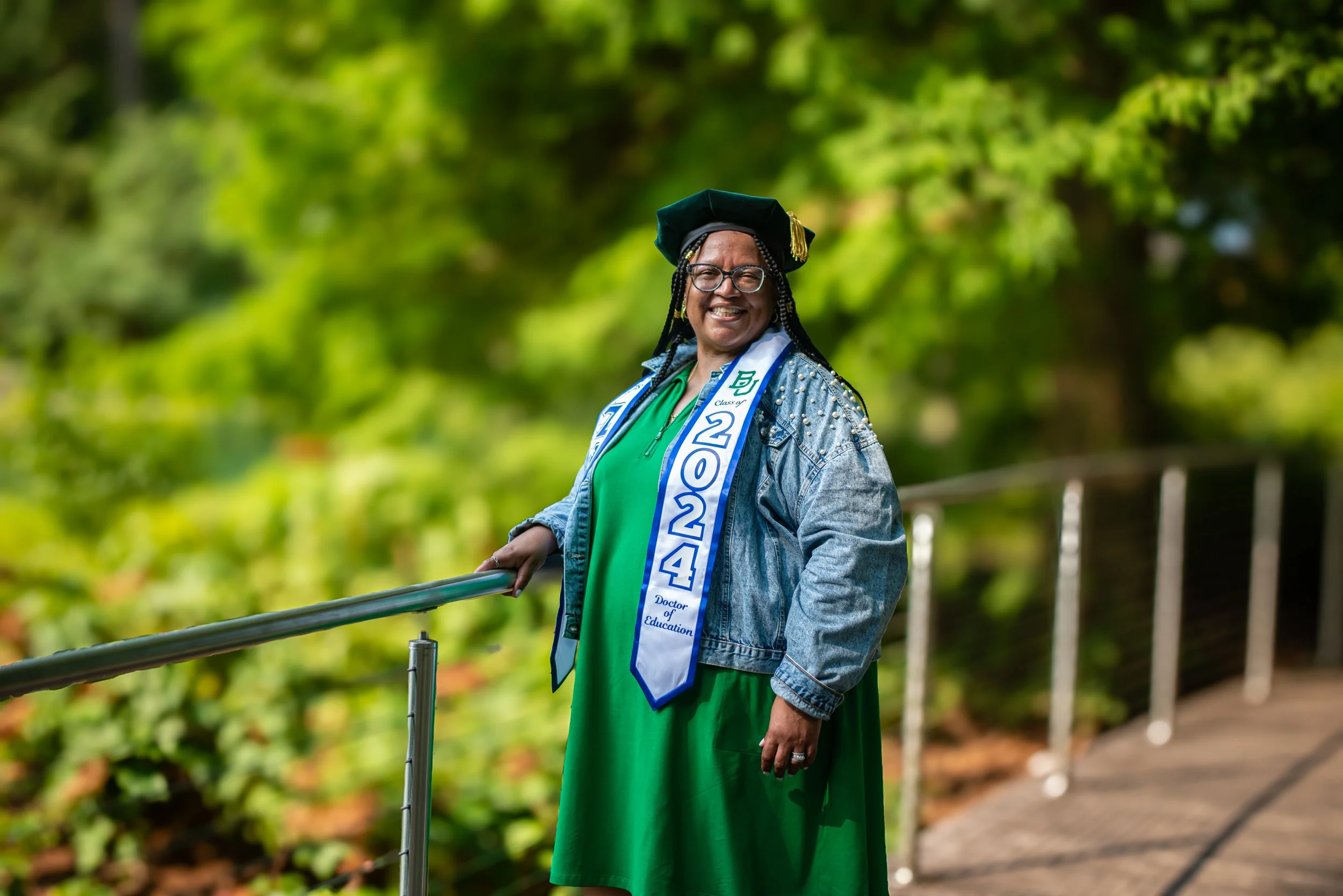 Woman dressed in green with a graduation cap and a sash that reads 'Class of 2024' stands outdoors smiling, with a green, leafy background.