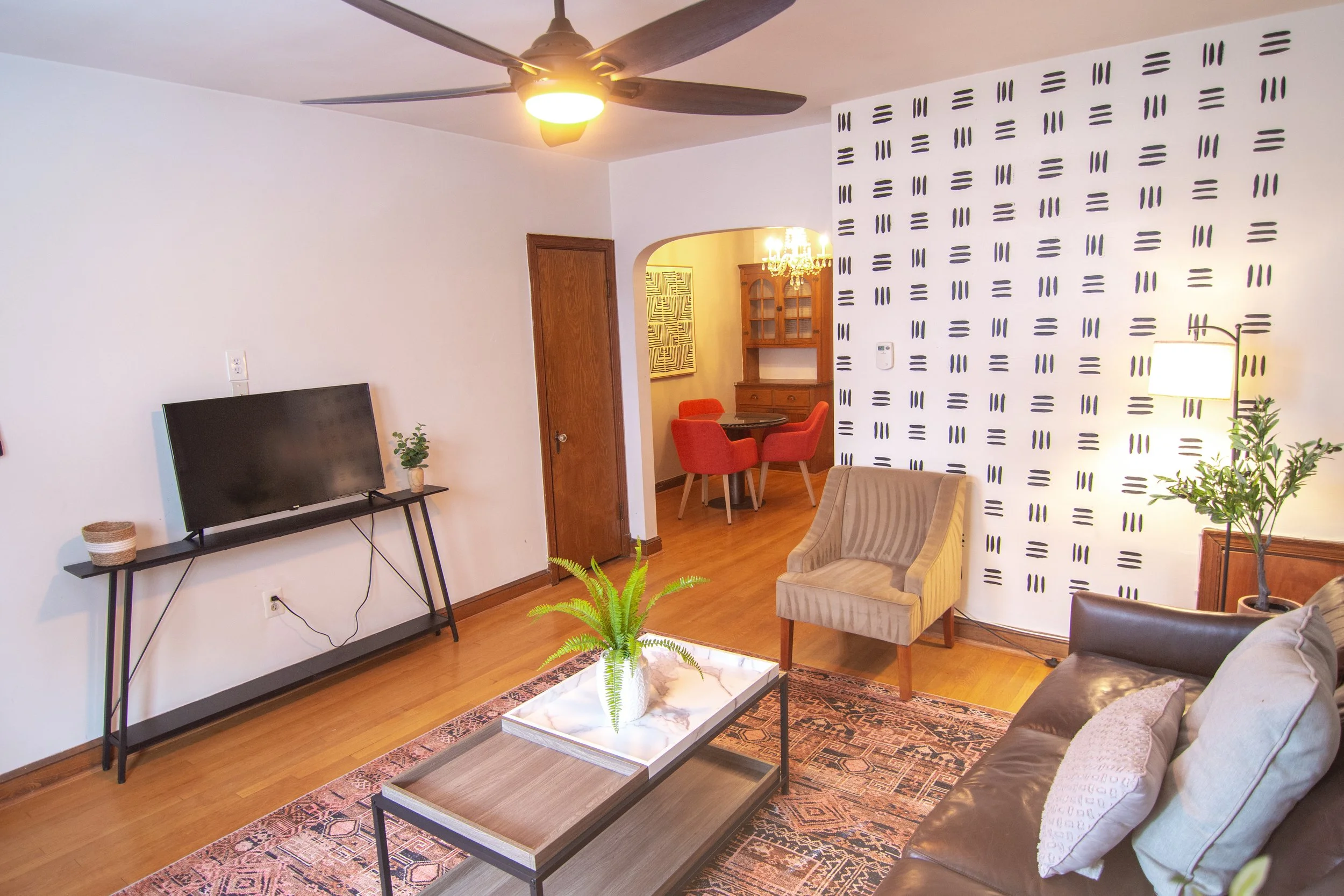 Living room with a flat-screen TV on a black stand, a beige armchair, a glass coffee table with a potted plant, a patterned rug, and a wooden dining area visible through an archway with a chandelier. A white wall with black geometric patterns and a c