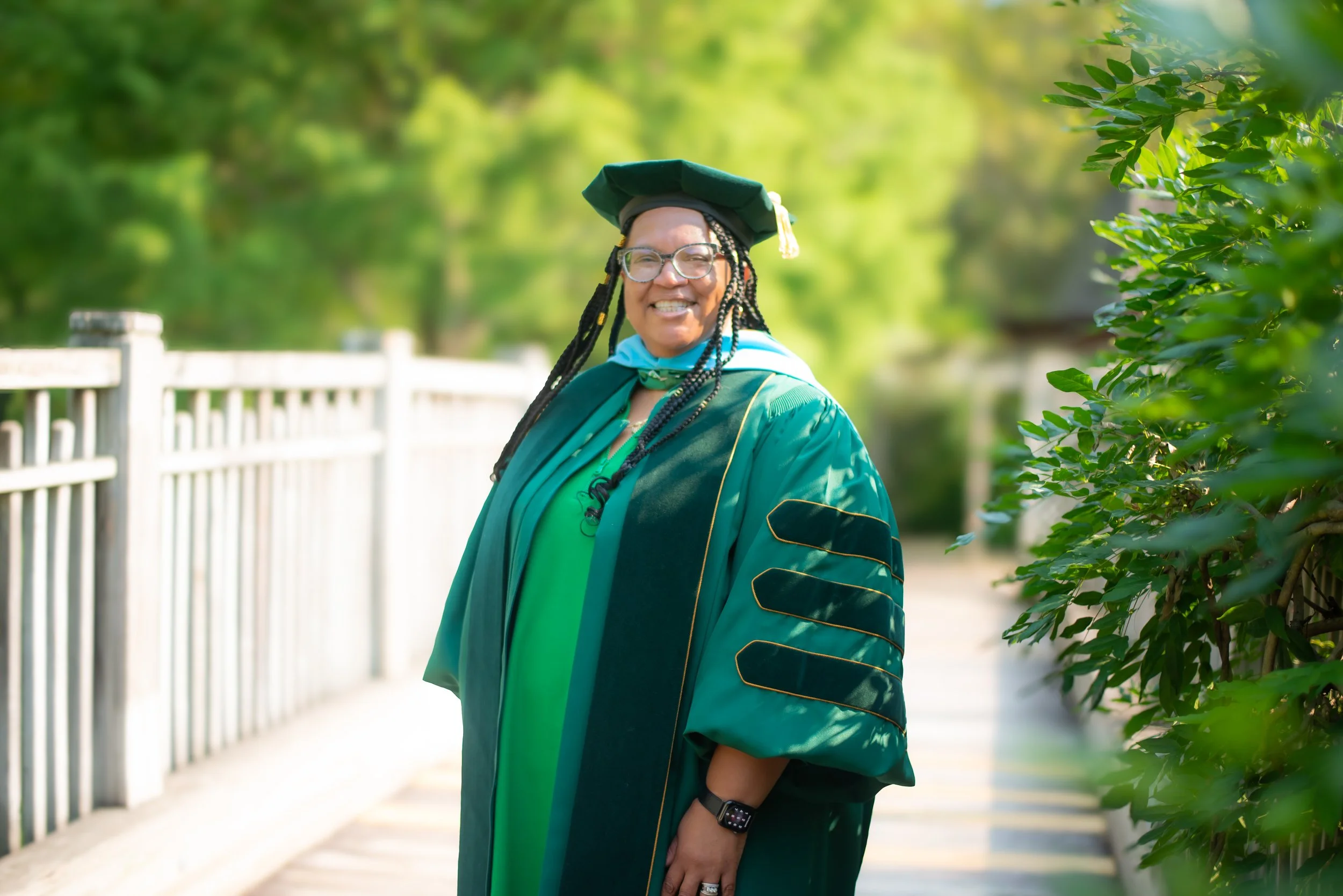 Woman in graduation cap and gown smiling outdoors with greenery in the background.