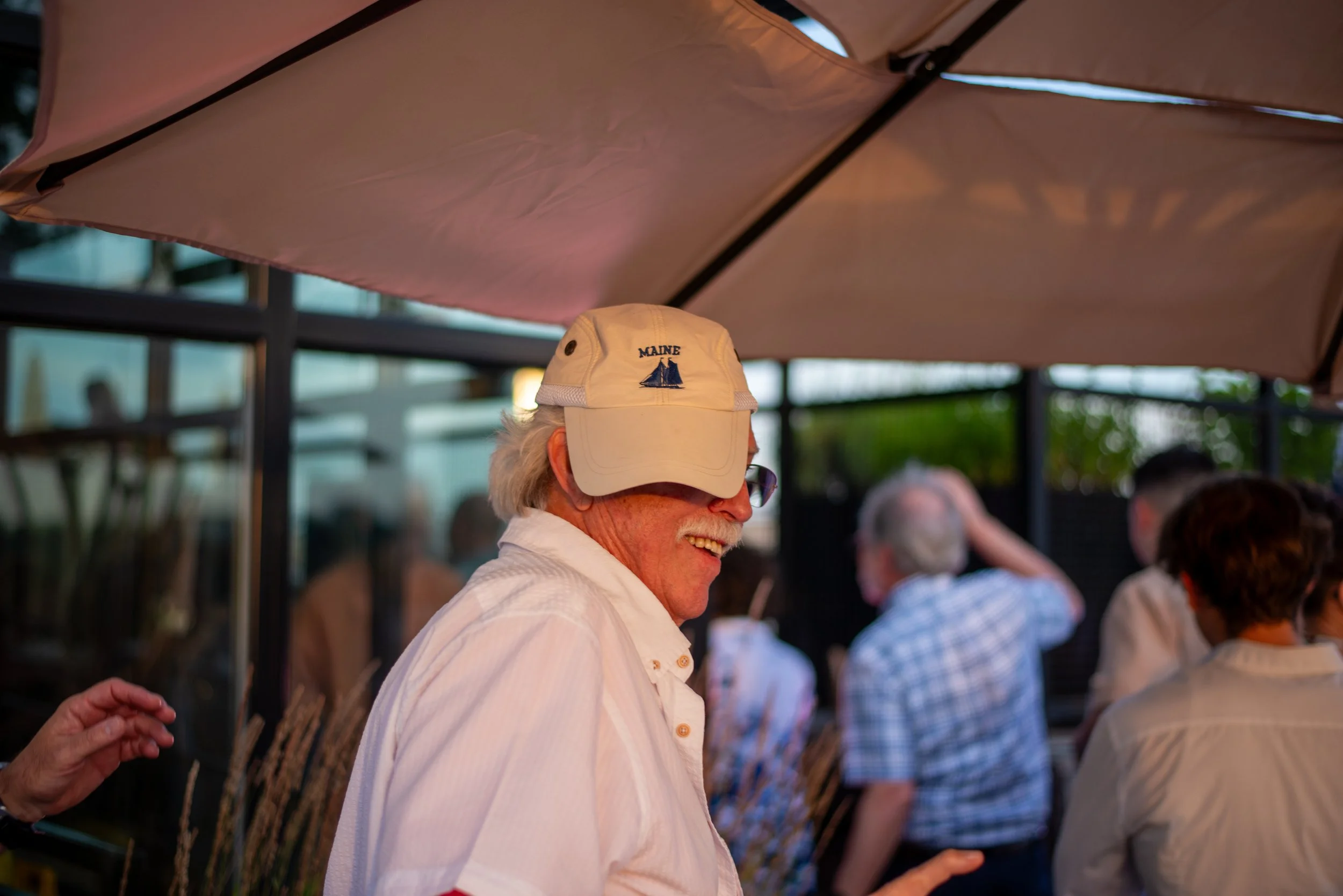 A man wearing a white cap with a sailboat logo and the word 'MAINE' on it, smiling at an outdoor gathering under an umbrella with other people visible in the background.