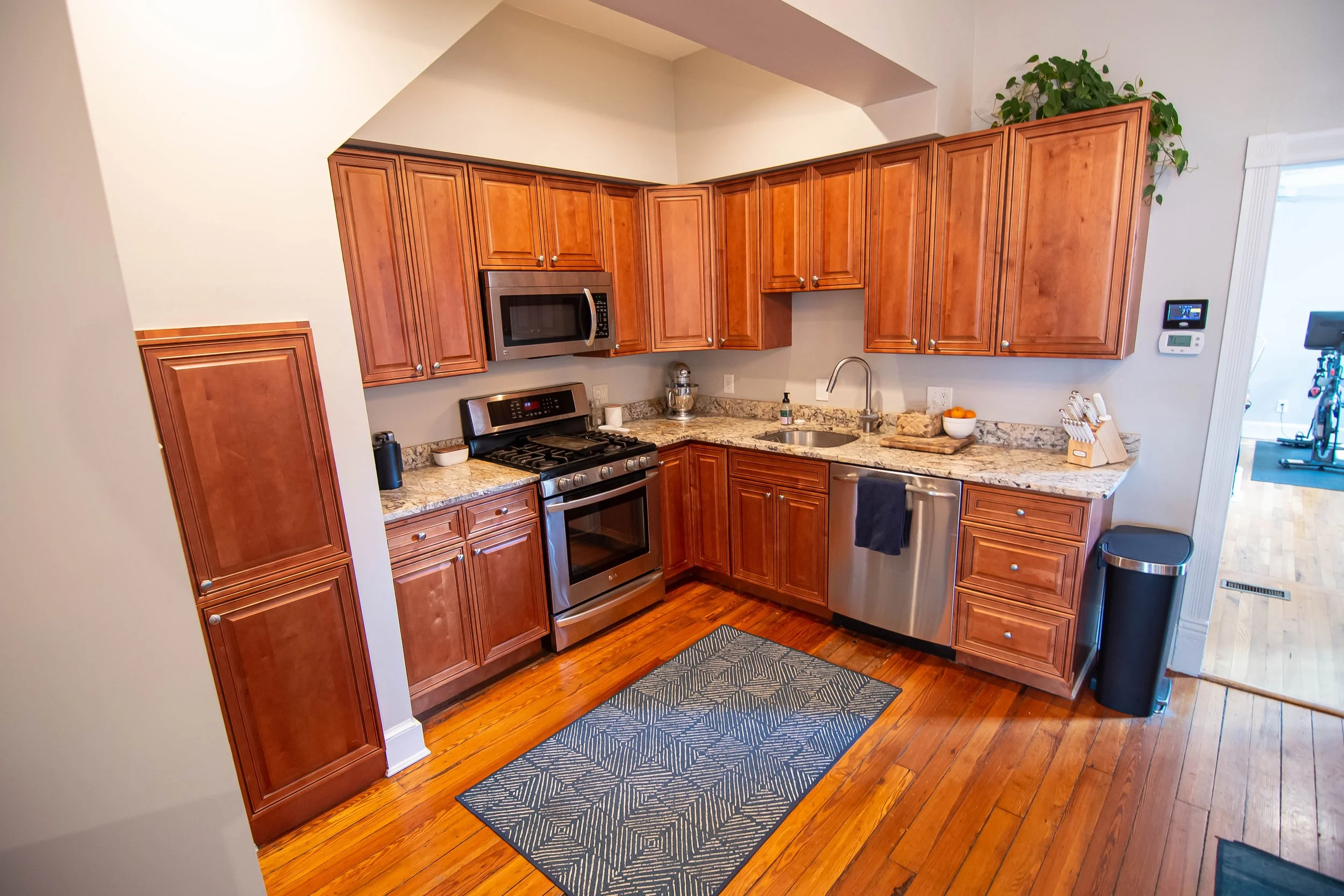 Kitchen with wooden cabinets, granite countertops, stainless steel microwave and oven, kitchen sink, small bowl of oranges, soap dispenser, and a black mat on polished hardwood floor.