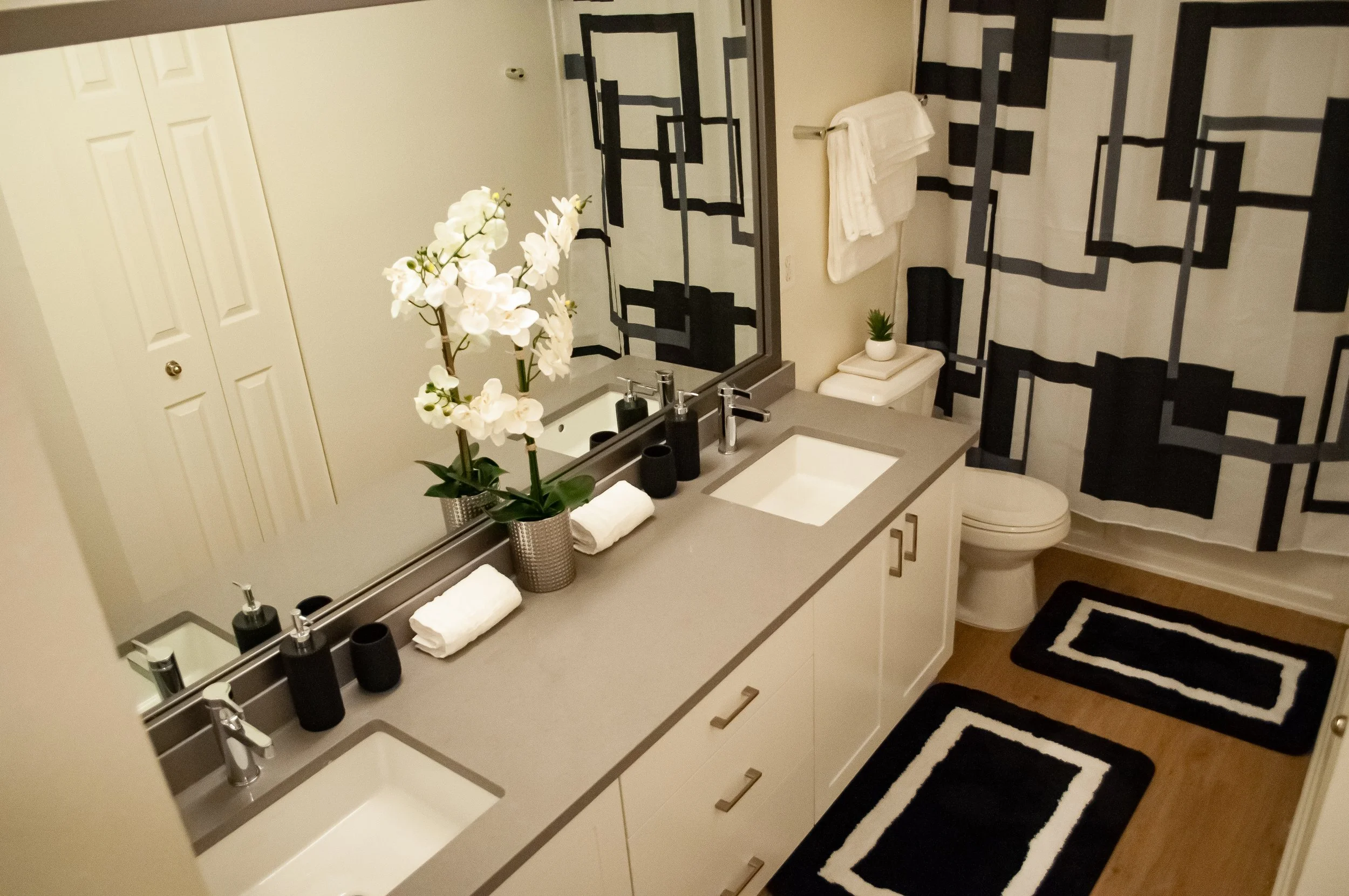 A modern bathroom with a double vanity, black and white geometric patterned shower curtain, a large mirror, and decorative items such as potted plants, towels, and soap dispensers.