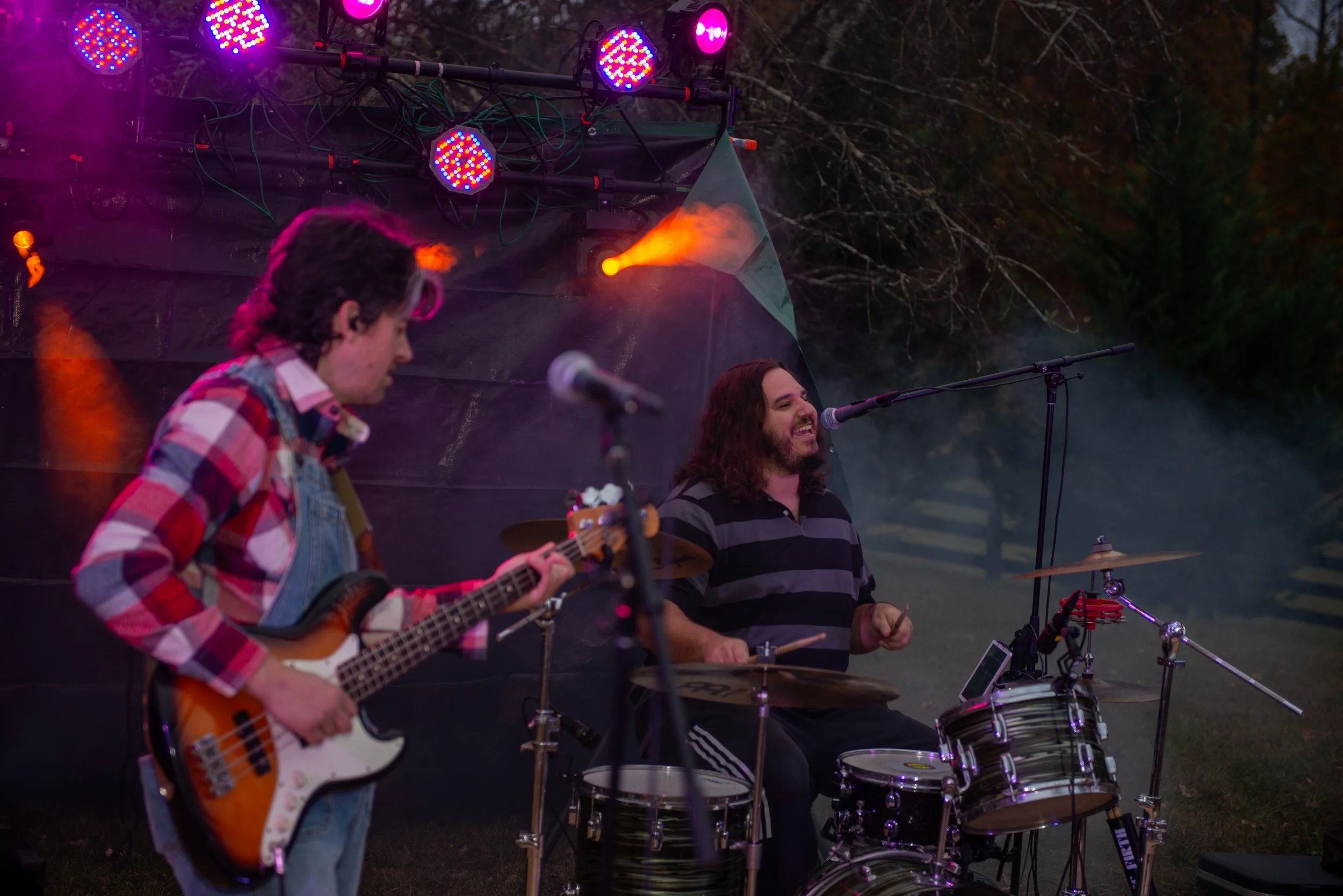 Two musicians perform outdoors at night; one playing an electric guitar and the other playing drums, with stage lights illuminating the scene.