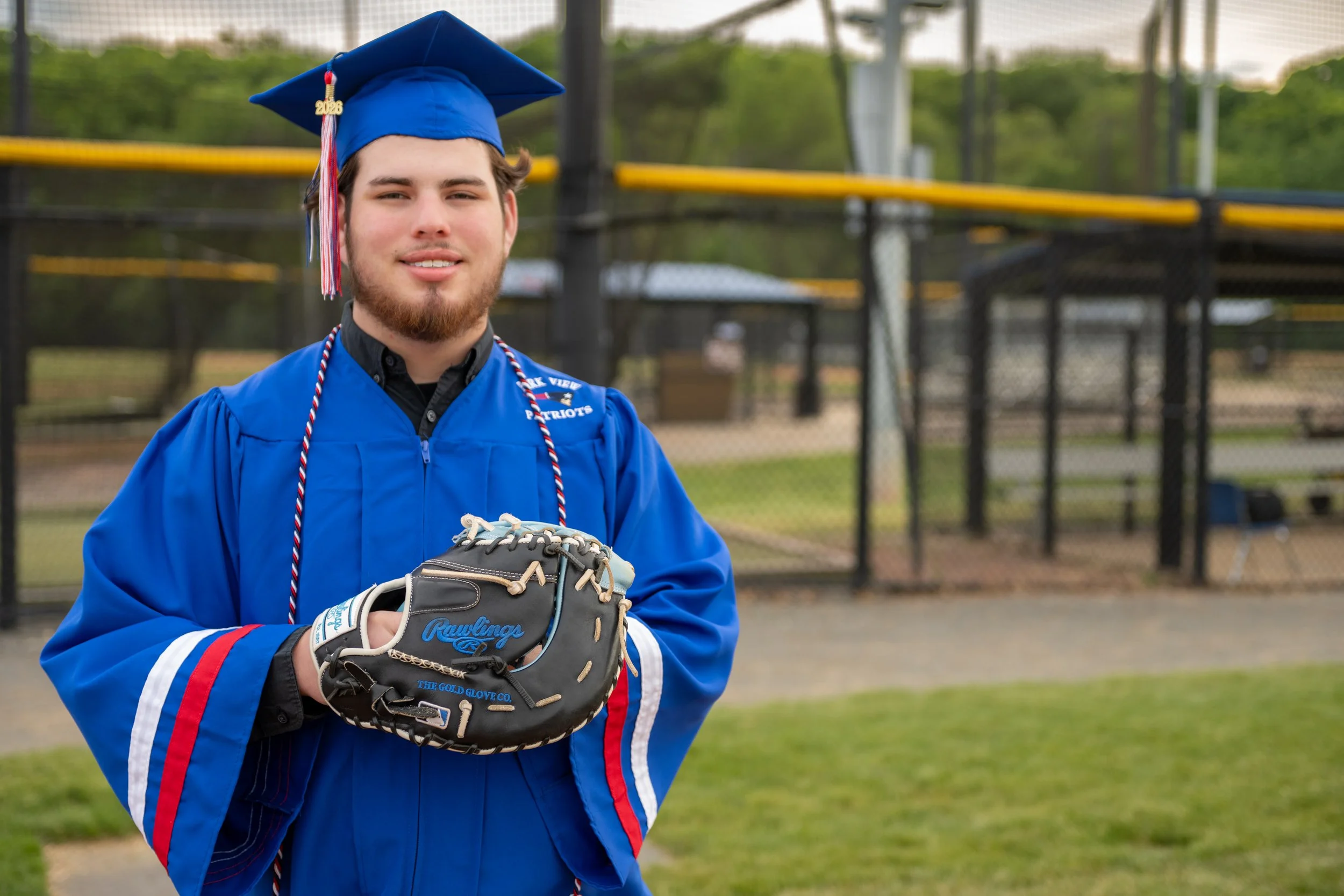 A young man in a blue baseball uniform and cap with a graduation year charm. He is holding a baseball glove labeled Rawlings and standing on a baseball field.