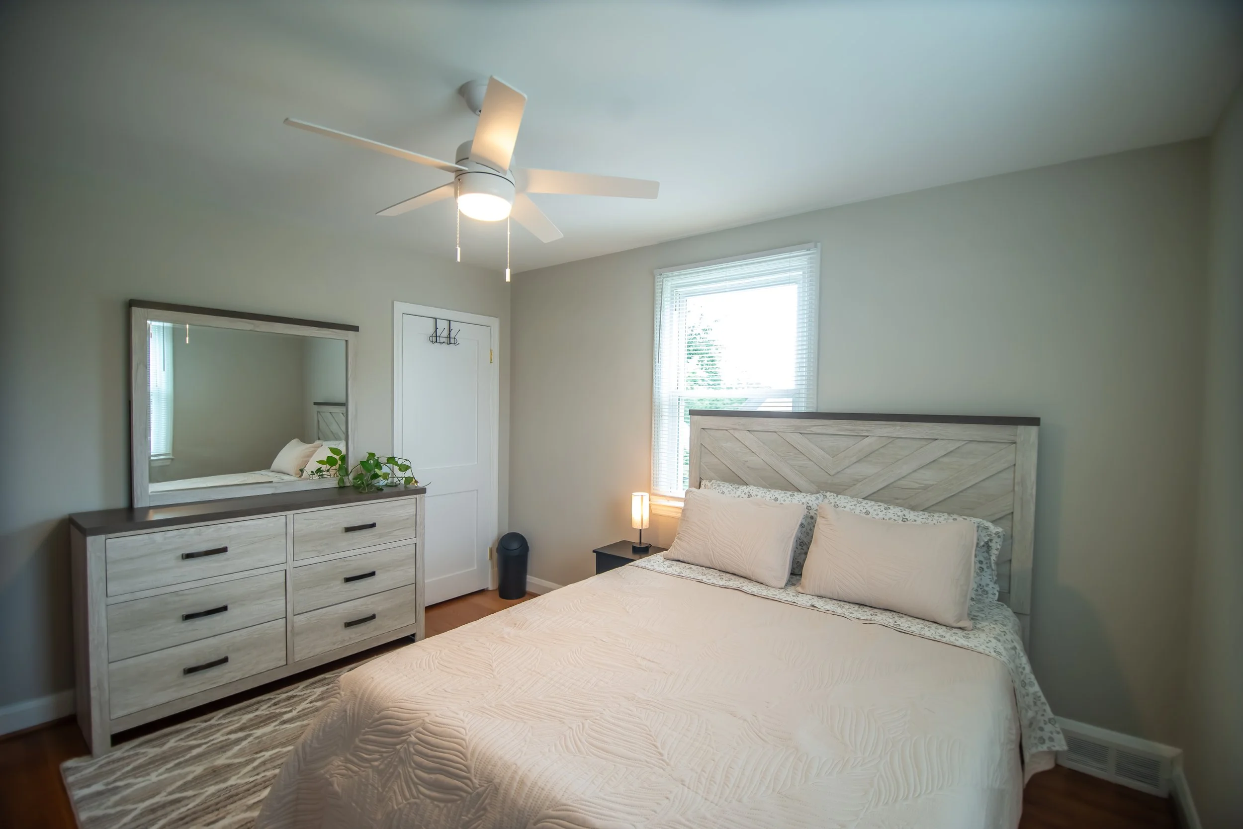 A bedroom with a bed, nightstand, dresser, and mirror, with a ceiling fan and window with blinds.