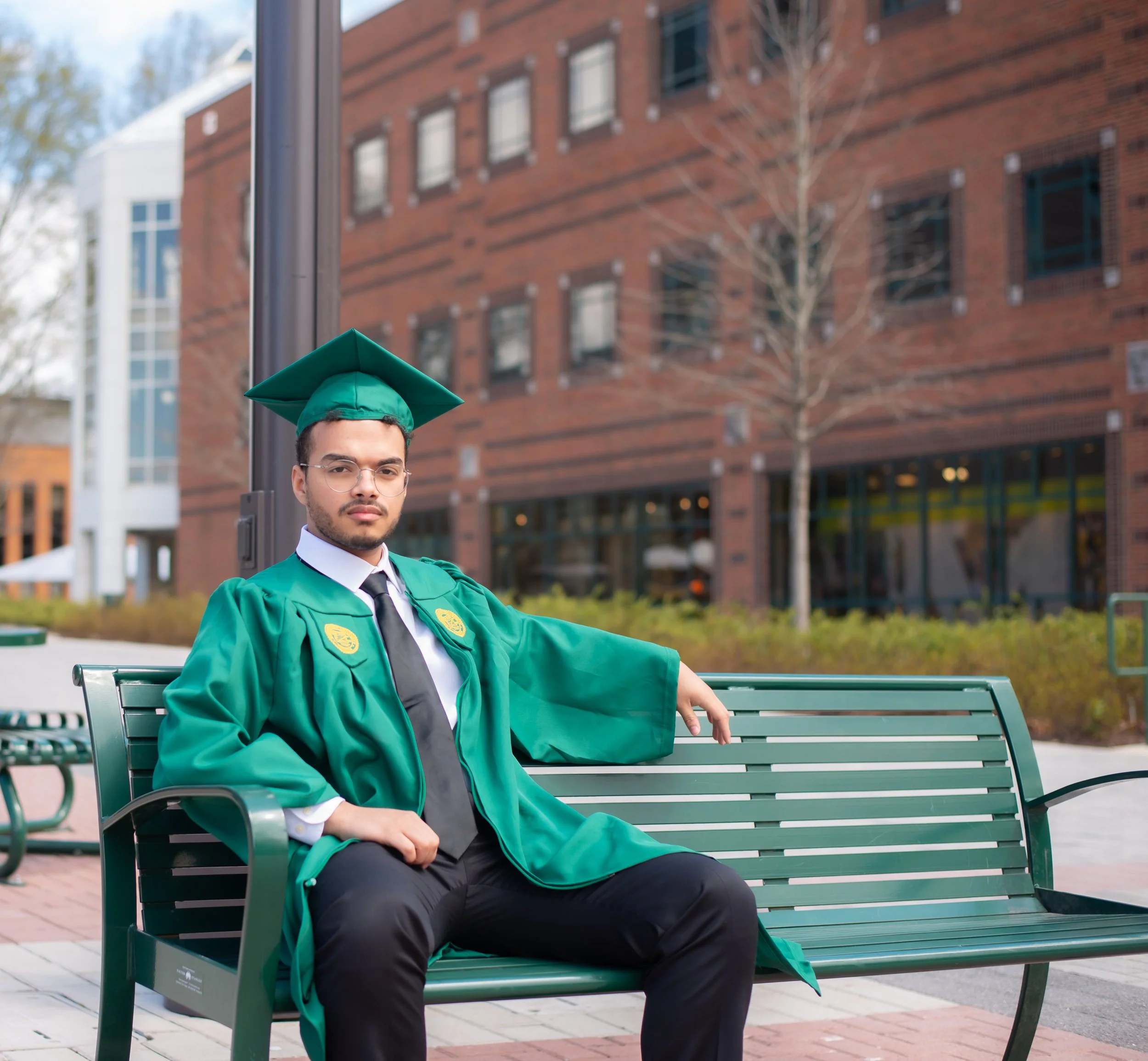 A man in a graduation cap and gown sitting on a green park bench.