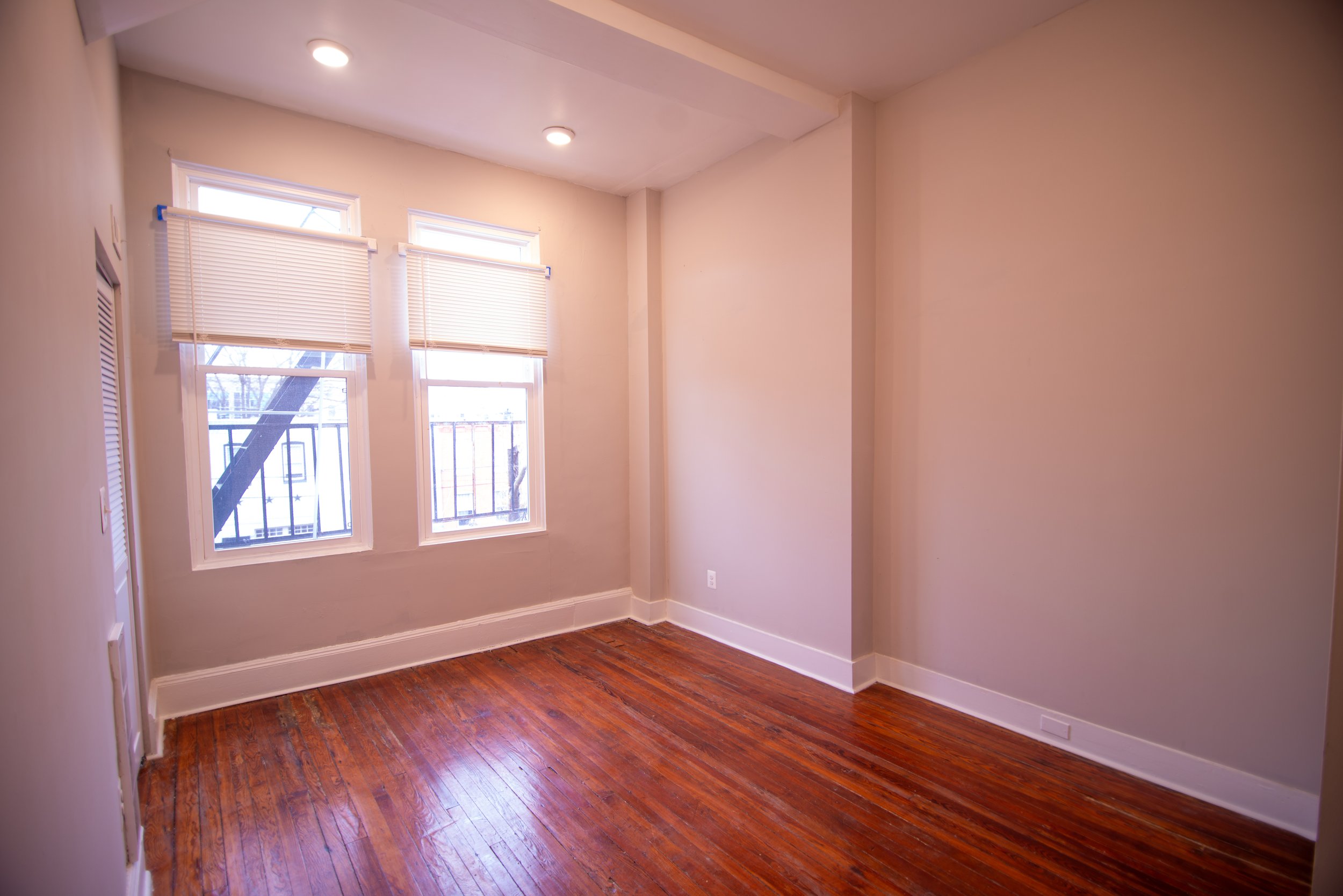 A small empty room with two windows with blinds, hardwood floors, and beige walls. There is a ceiling light turned on and an electrical outlet near the floor.