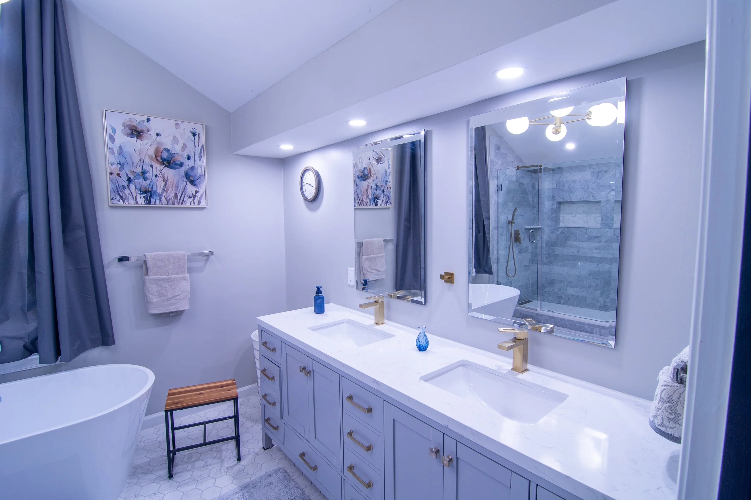 Modern bathroom with white vanity, large mirror, and walk-in shower with glass door and grey tiled walls. Decorative art and clocks on walls, with towel racks, a white bathtub, and a stool beside the tub.