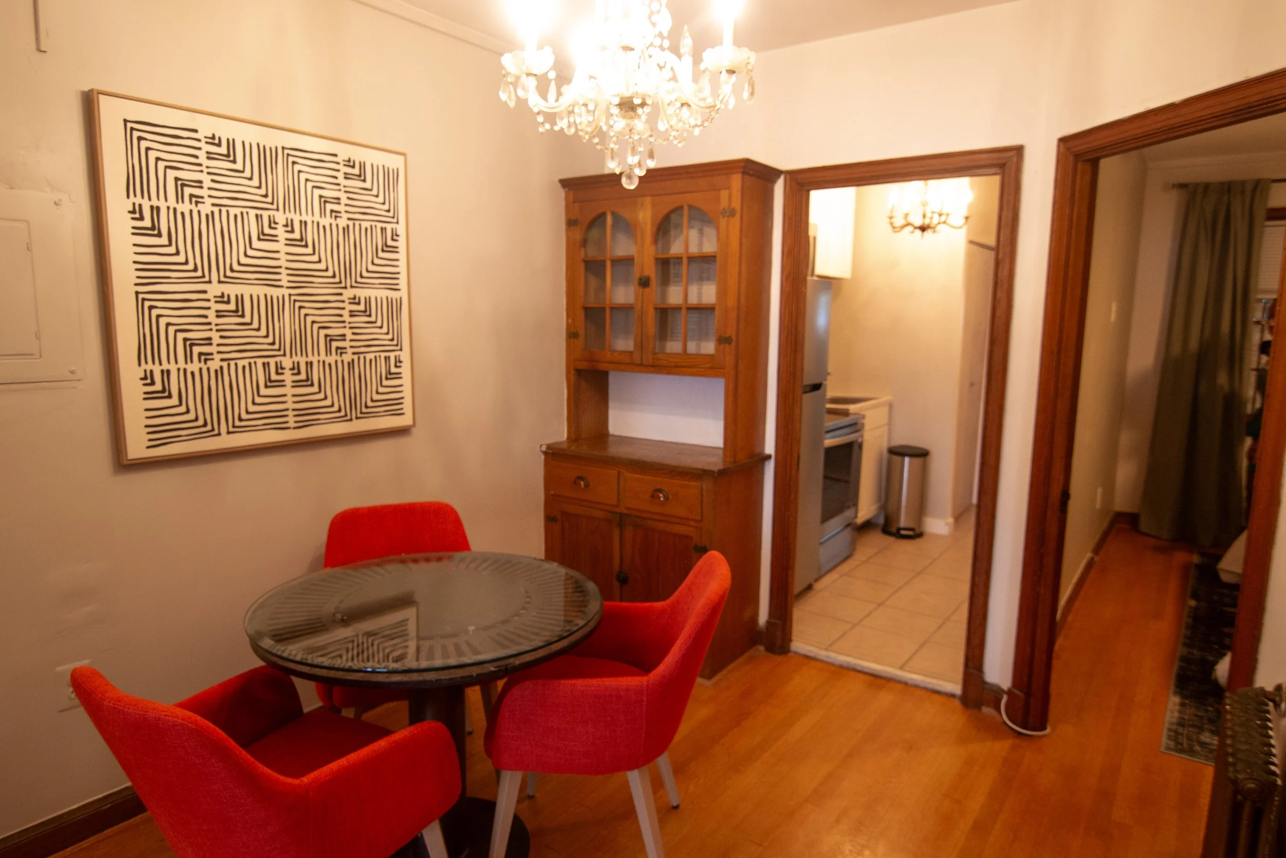 A dining area with a small round glass table surrounded by four red upholstered chairs. An abstract black and white geometric art piece is on the beige wall. In the background, there is a wooden cabinet with glass doors and a kitchen area with a stov