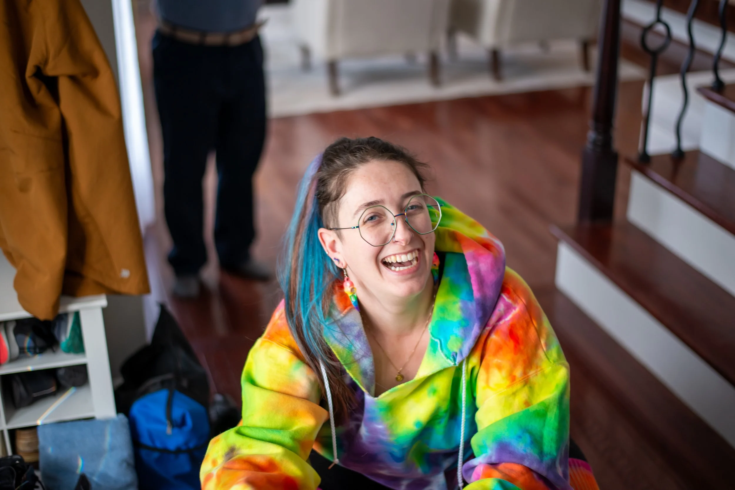 A person with glasses and colorful tie-dye clothing is smiling and looking up at the camera. There is a person in the background wearing a blue shirt and another on the left in a yellow jacket, with some household items visible around.