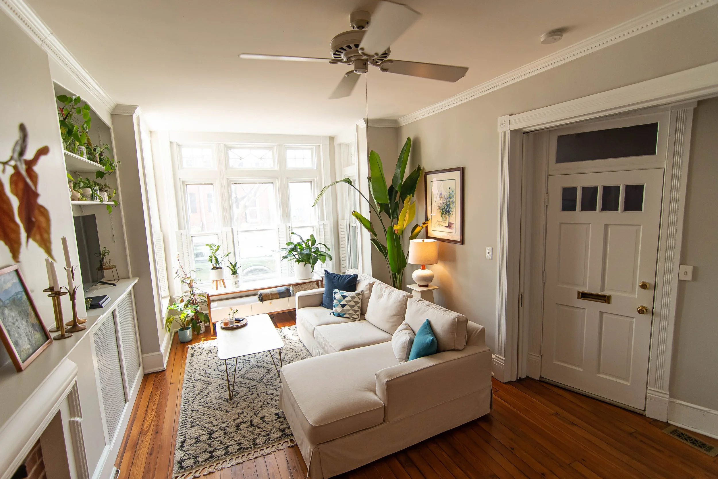 A living room with a large bay window, white walls, and hardwood floors. There is a white sofa with colorful pillows, a wooden coffee table with a tray, and various indoor plants. A framed artwork hangs on the wall, and a ceiling fan is visible.