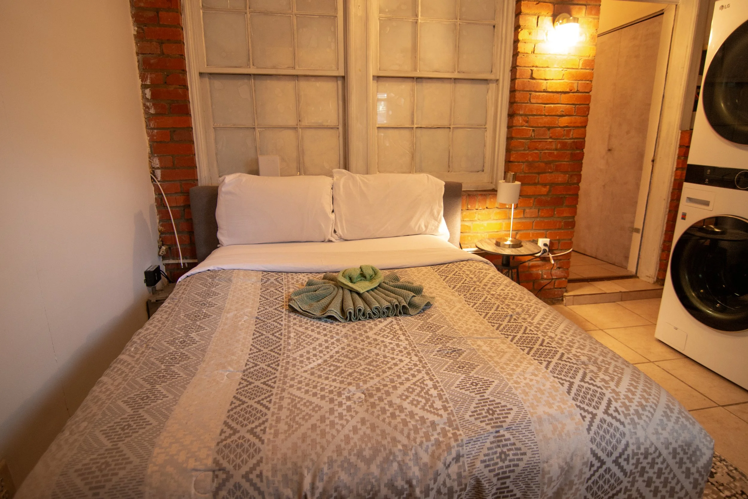 A cozy bedroom with a bed covered in a beige geometric patterned quilt, featuring two white pillows, and a neatly folded green towel on top. The headboard is against a brick wall, with large old windows above, and a small round bedside table with a l