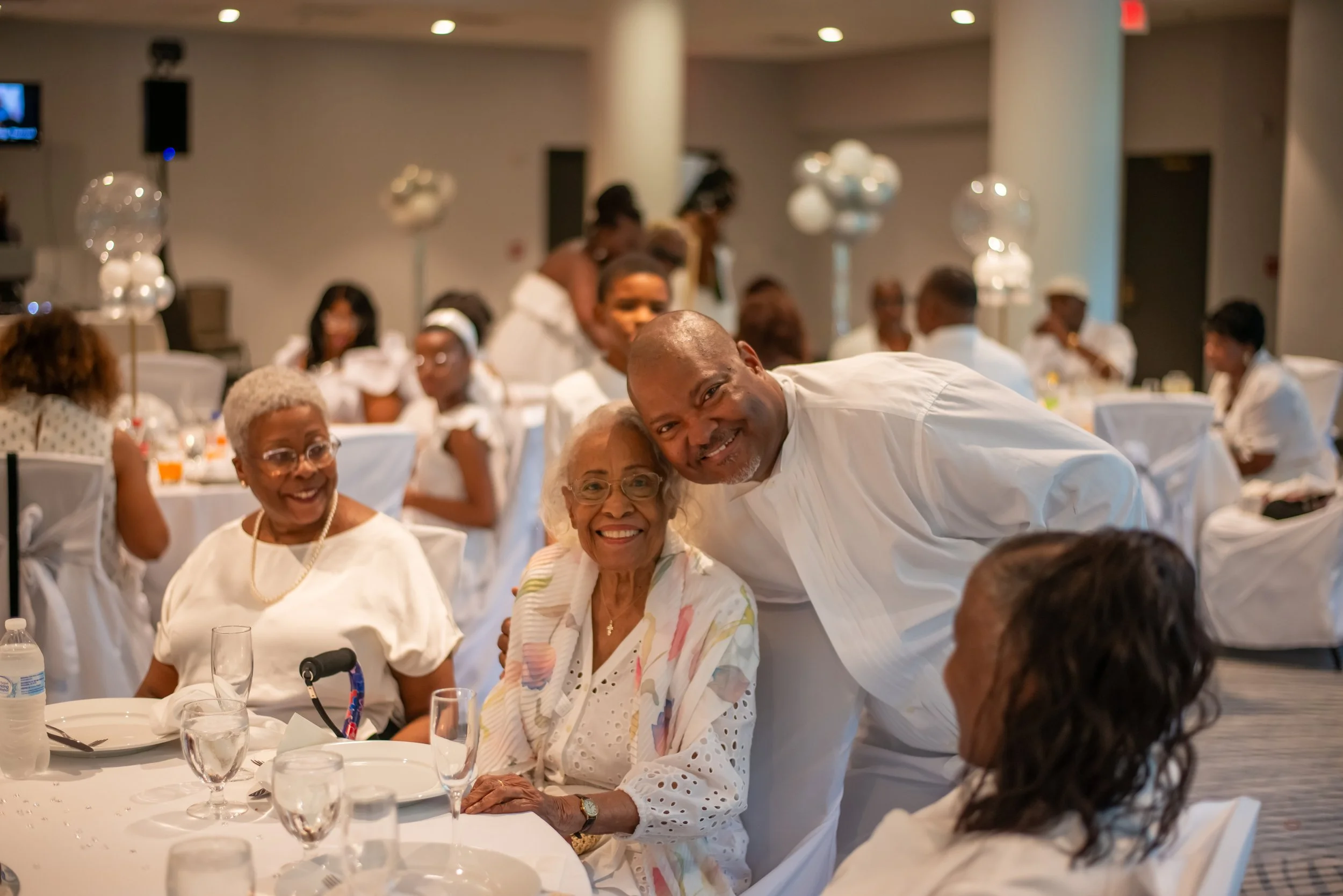 A group of elderly and middle-aged people gathered at a banquet, smiling, with a man leaning over to a seated woman in focus. The scene appears to be festive, with decorated tables and balloons.