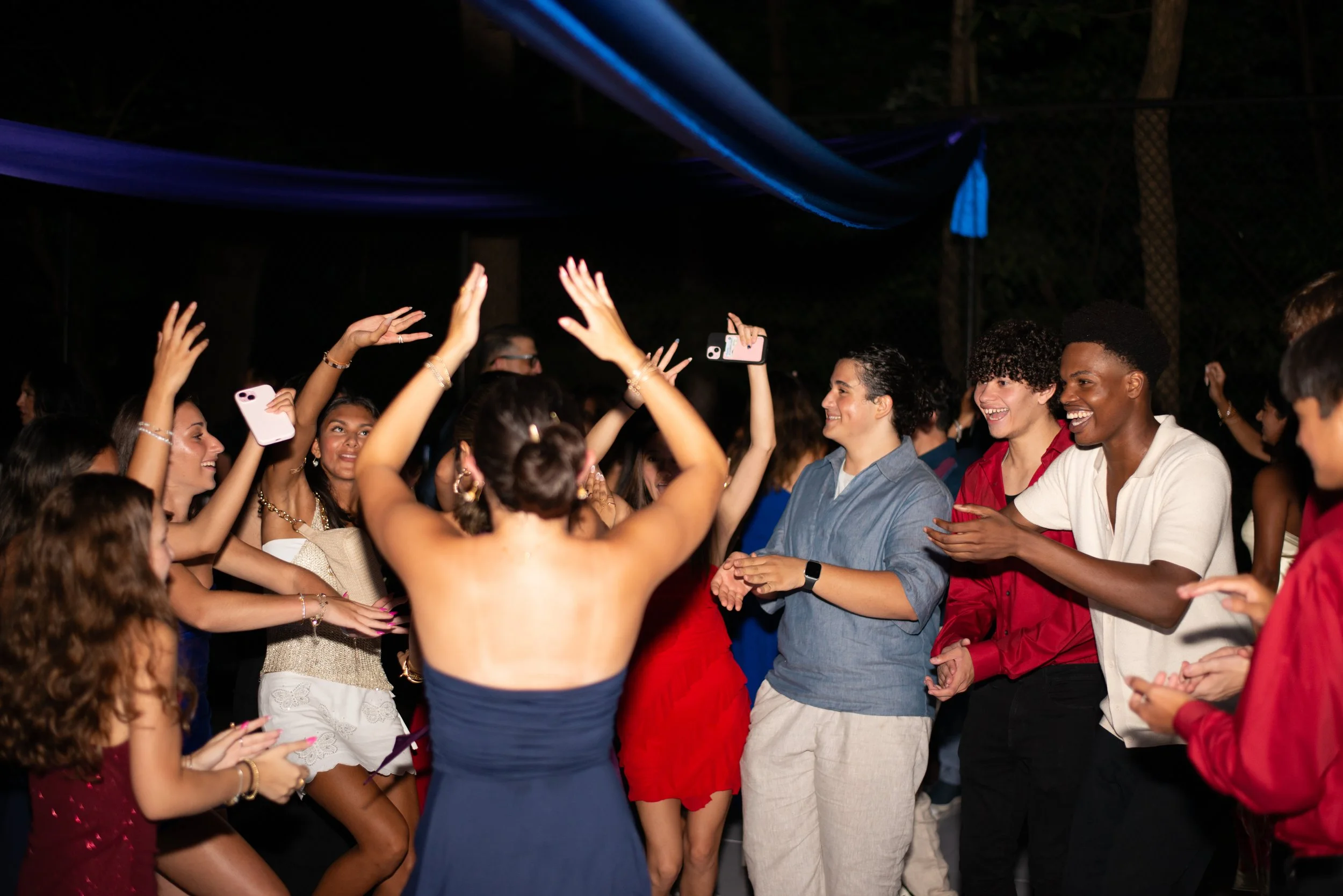 People dancing and celebrating at a lively party or event outdoors at night, with some holding phones to take photos or videos.