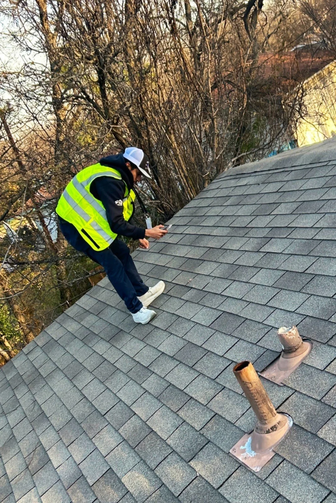 Roofer in a safety vest checking asphalt shingles near two pipe vents during a hail damage roof inspection