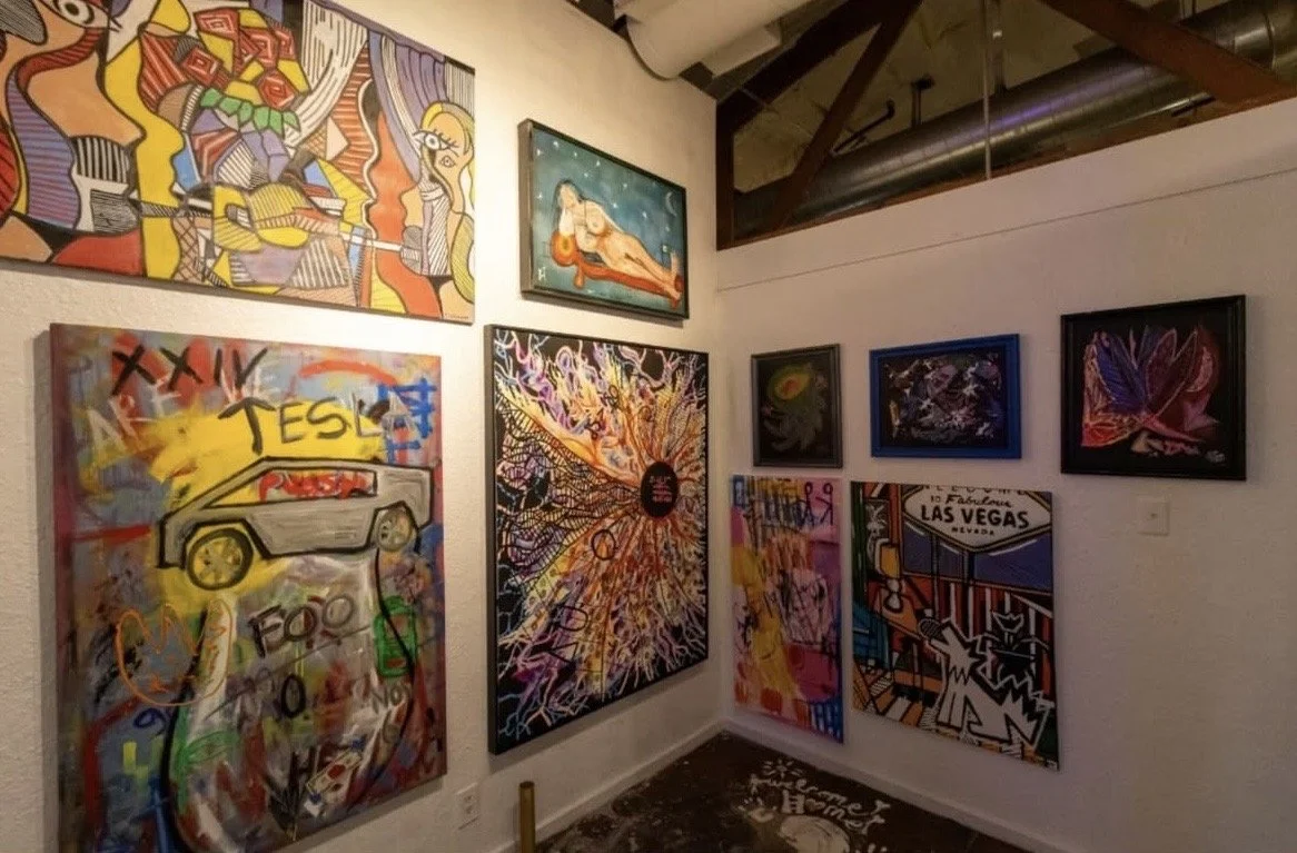 Gallery wall featuring colorful abstract and pop art paintings, including a Tesla-themed artwork, a reclining nude, and a Las Vegas sign, in an indoor art gallery space with a high ceiling.