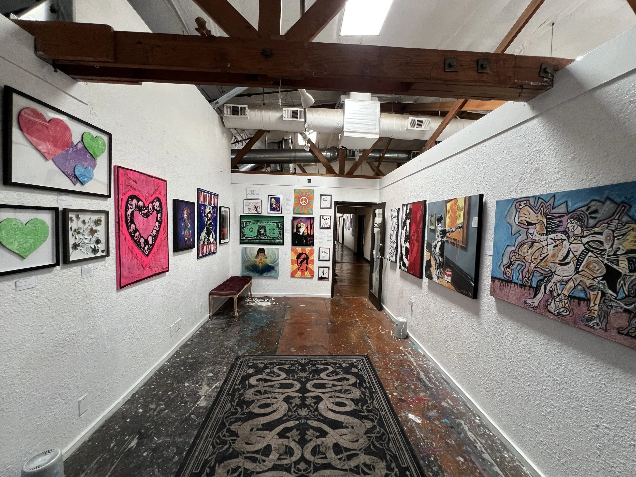 Interior view of an art gallery with white textured walls and wooden ceiling beams. Artworks include colorful hearts, a money-themed piece, portraits, and abstract paintings. There is a black and gold patterned rug on the floor and a small bench along the left wall. The gallery space appears to have natural light coming through a skylight.