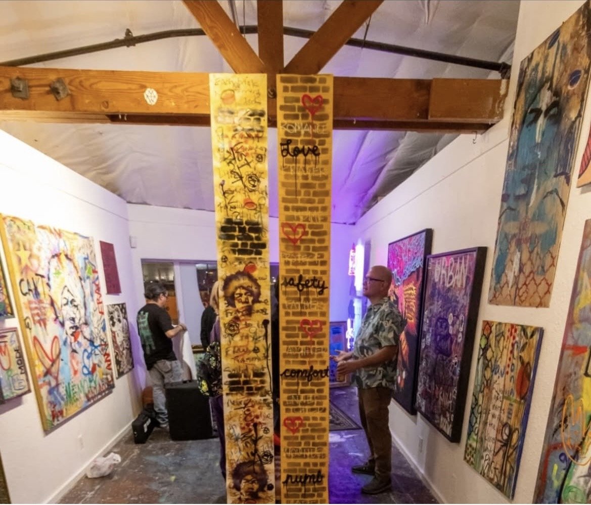 Art gallery with paintings on walls and graffiti-style murals on hanging panels, wooden beams on ceiling, people observing artwork.