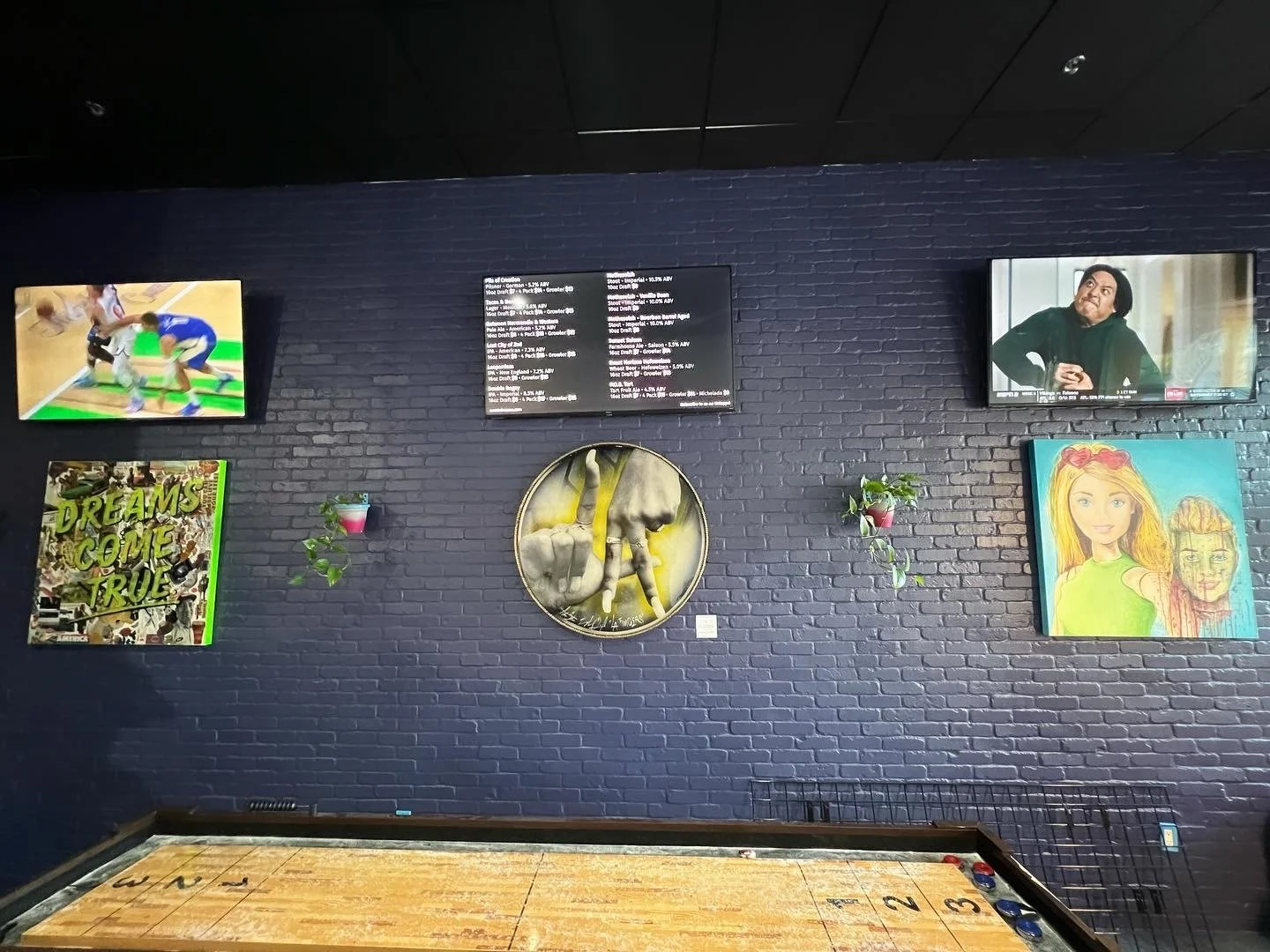 Black brick wall with five mounted screens and artworks, and a shuffleboard table at the bottom.