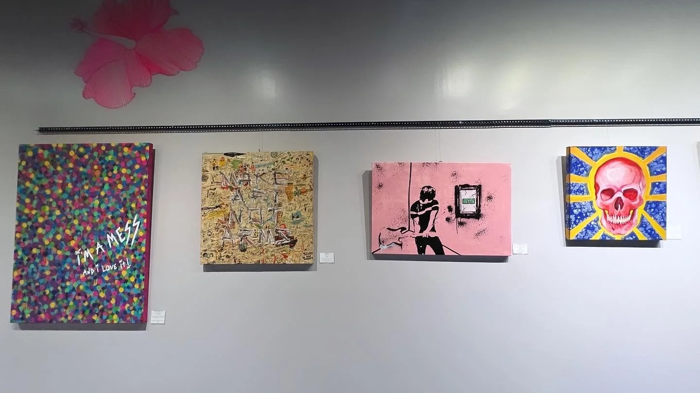 Gallery wall with four colorful paintings, including a multicolored dot pattern with text 'I'M A MESS AND I LOVE IT!', abstract pieces, a stencil of a boy sitting with a dog on pink background, and a skull with sun rays.