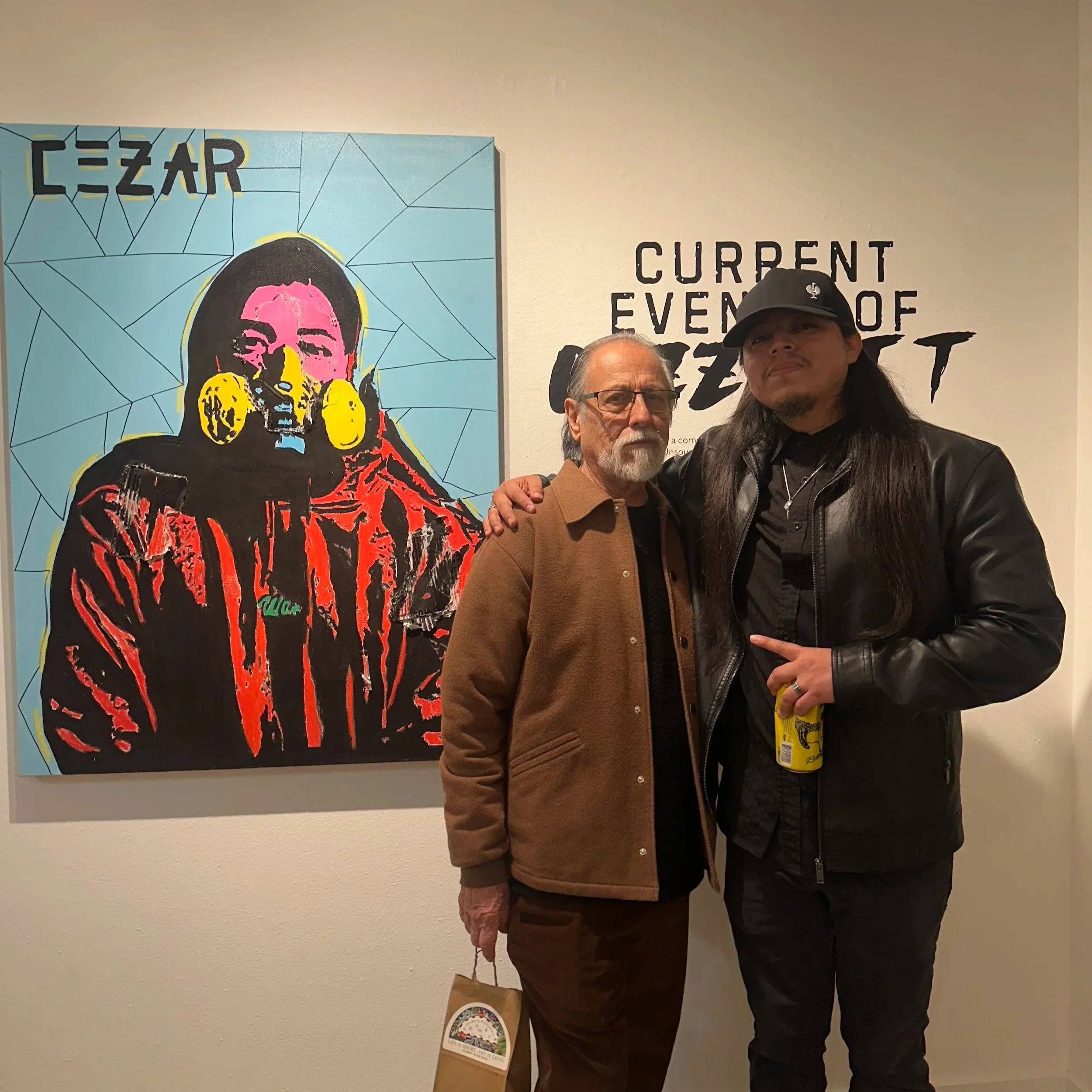 Two men standing together in front of a colorful art piece at an exhibit. One is an older man with glasses, a beard, and wearing a brown jacket. The other is a younger man with long hair, wearing a black leather jacket and a black cap, holding a beverage can.