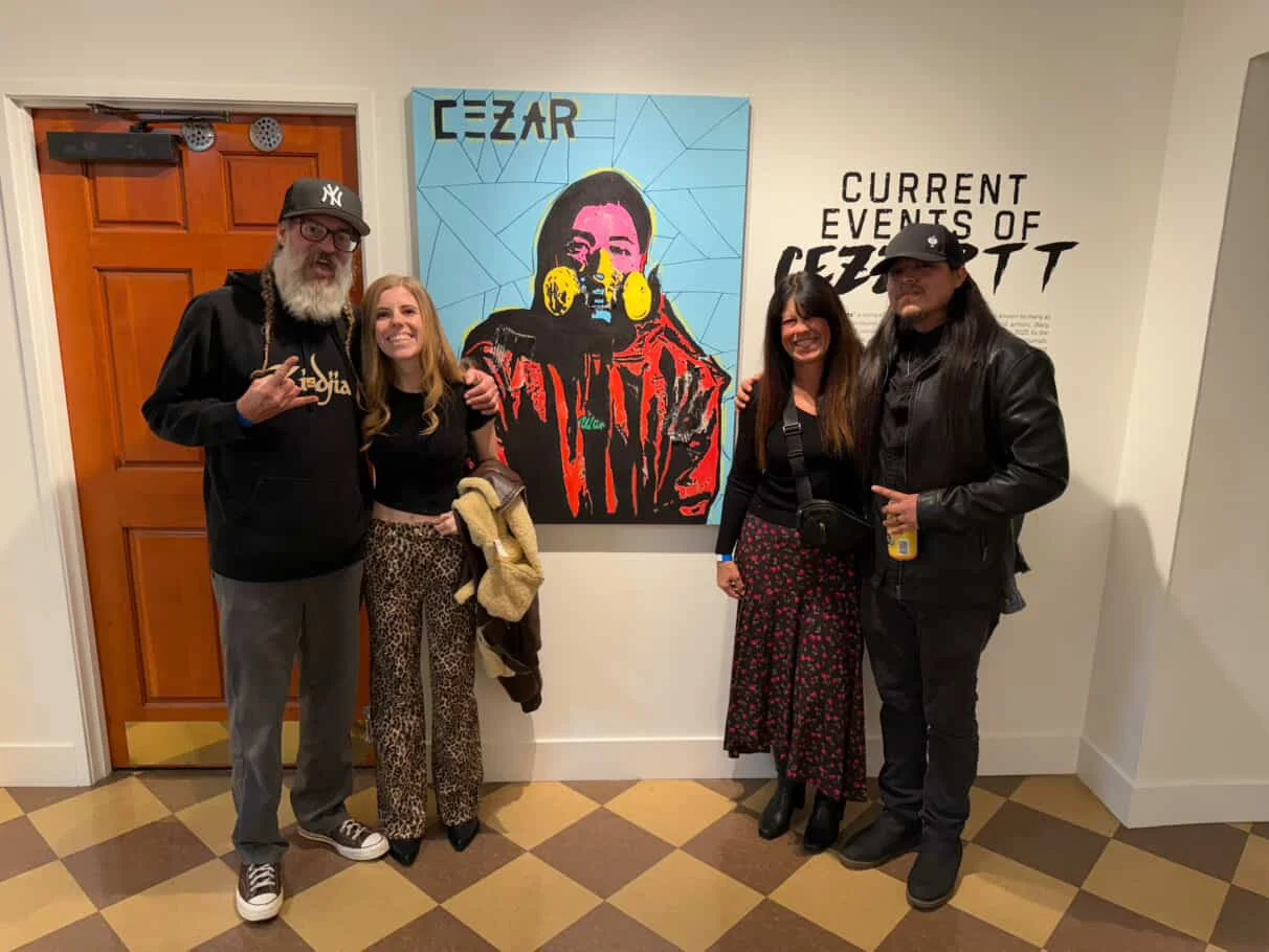 Four people standing in front of a colorful portrait CEZZARTT, with text indicating an exhibition. The two men are wearing dark clothing and hats, and the two women are smiling, one holding a jacket.