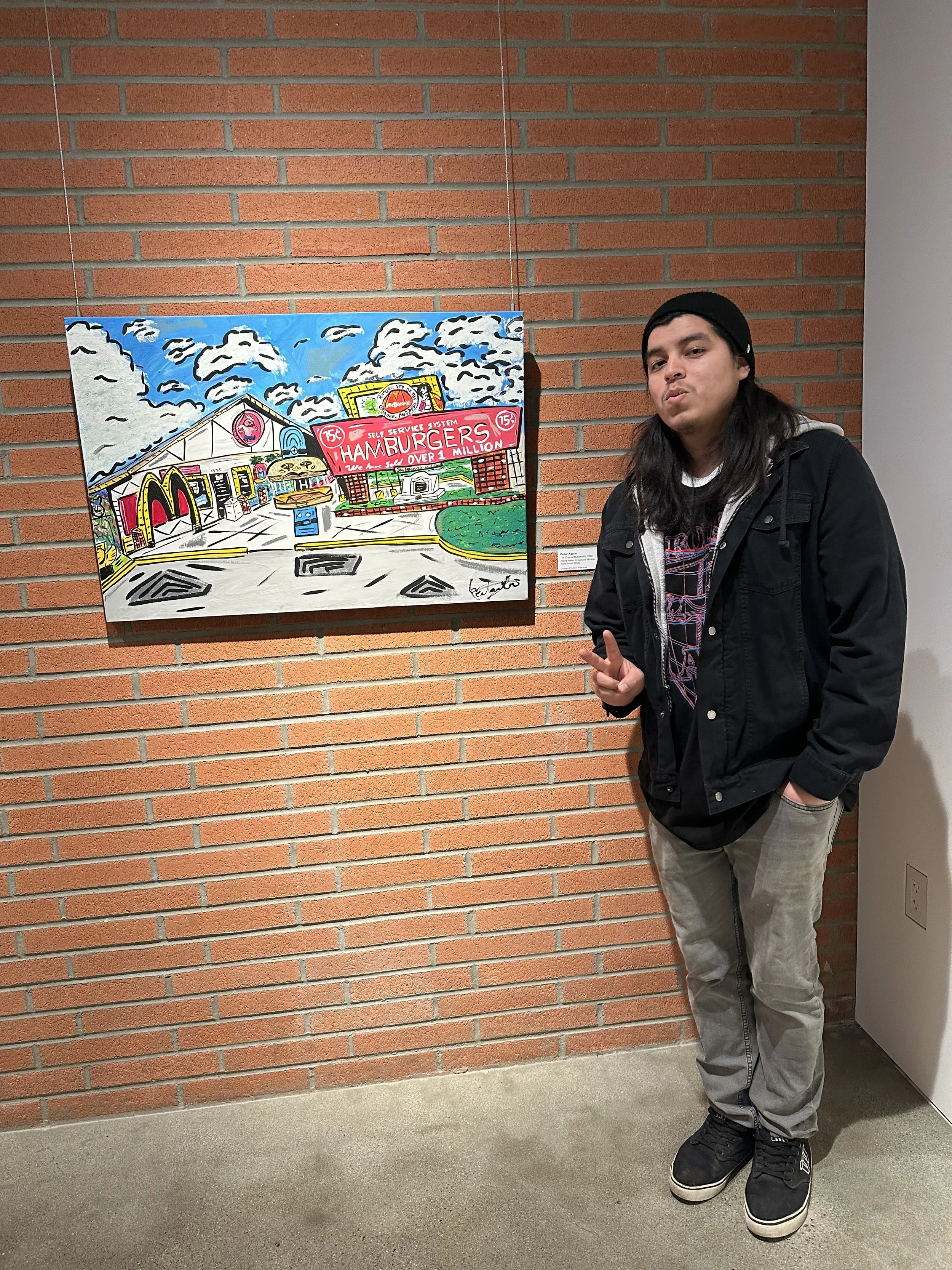 A person with long dark hair, wearing a black jacket, gray pants, and a black beanie, standing next to a colorful painting of a McDonald's restaurant on a brick wall.