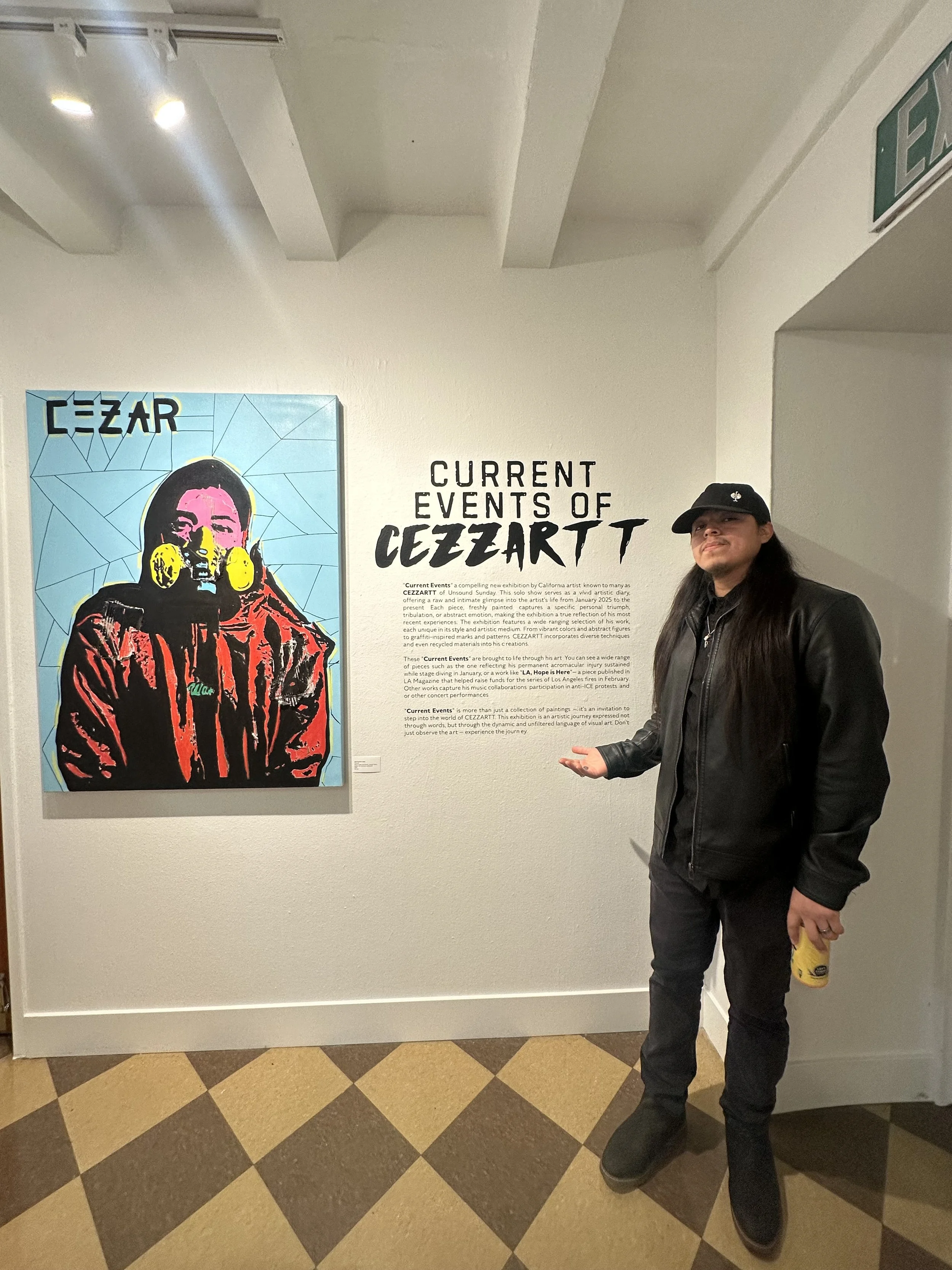 A person standing in front of an art exhibit titled 'Current Events of CEEZARTT' with a colorful portrait of a woman wearing headphones, in an art gallery.