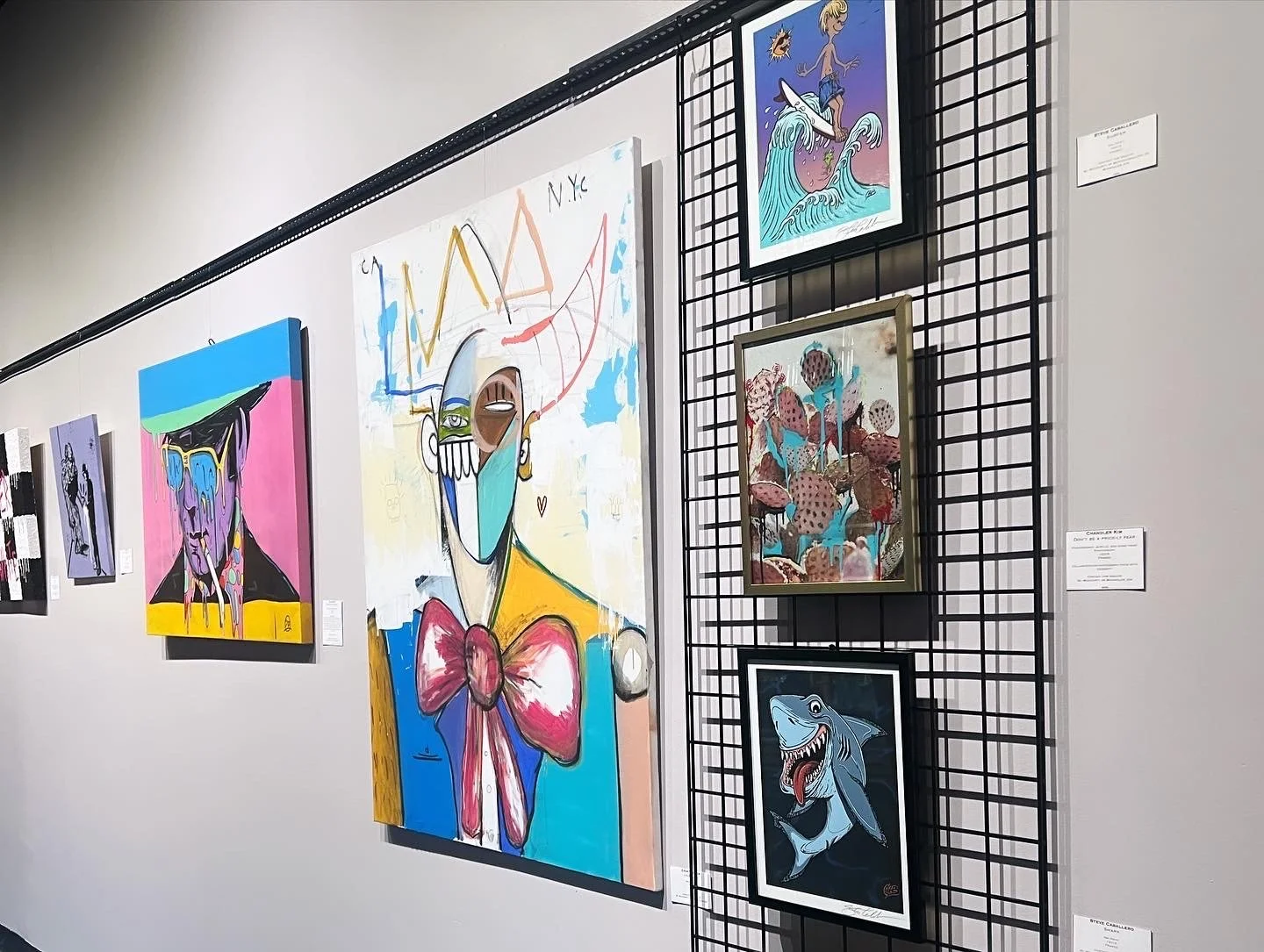 Gallery wall displaying various colorful artworks, including abstract portraits, a surfing figure, a fish, and scenes with abstract elements, arranged on a white wall with one artwork mounted on a black grid.